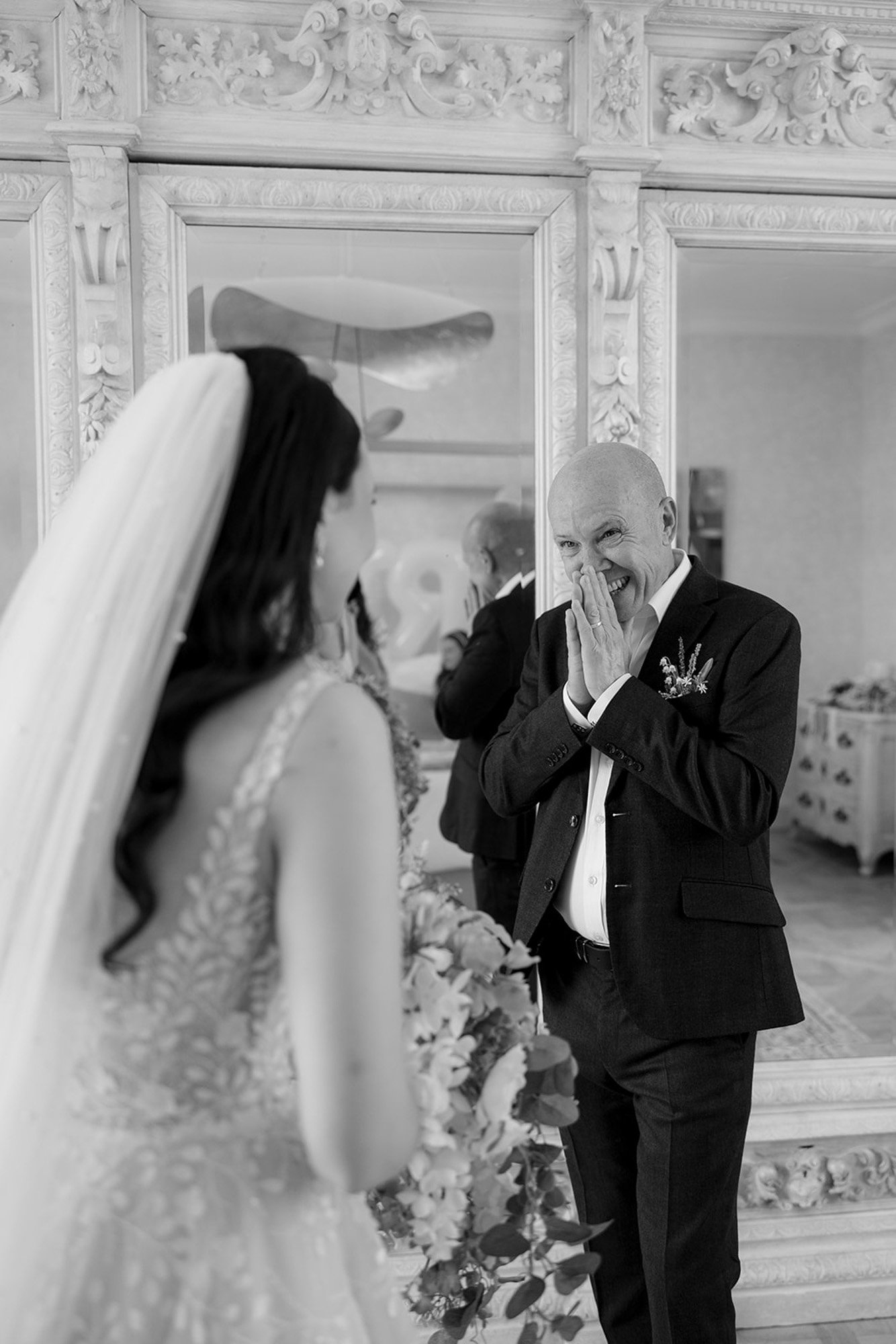 A bride stands facing an older man in a suit, who appears emotional with his hands covering his mouth, in front of a large ornate mirror. Spring Dordogne Wedding Photographer