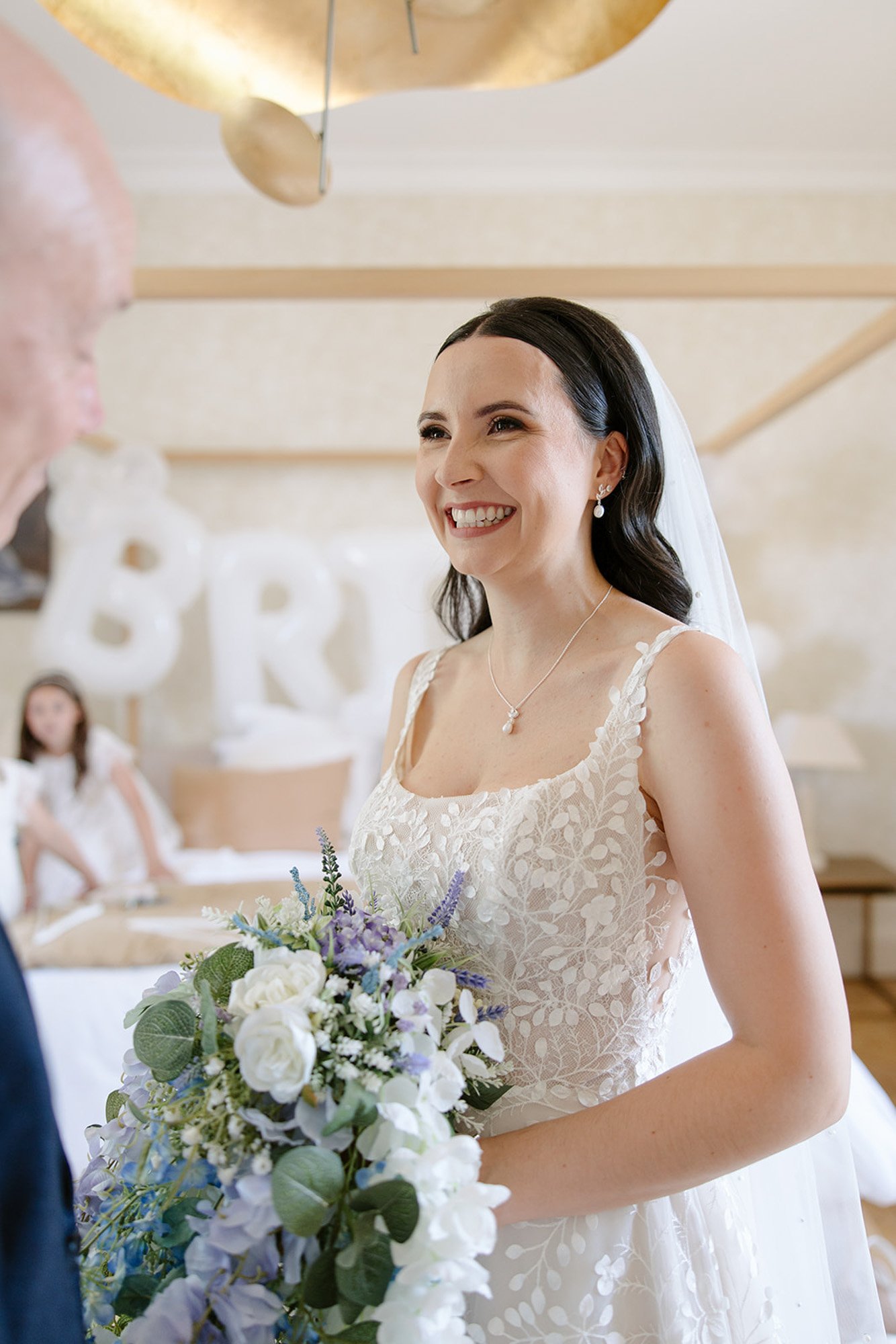 A bride in a white lace dress and veil holds a bouquet of white and purple flowers, smiling indoors with blurred BRIDE balloons in the background.