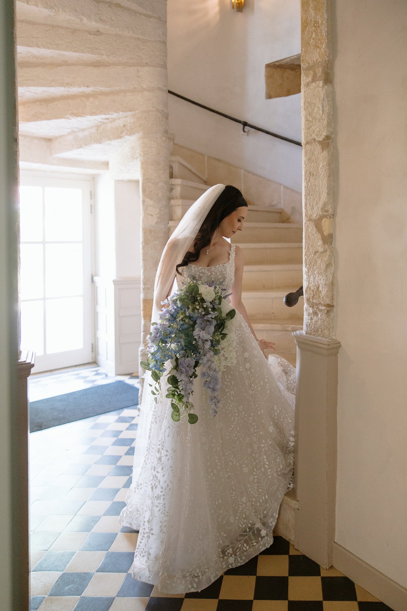 A bride in a white lace wedding dress and veil holds a large bouquet of blue and white flowers while standing by a stone staircase indoors. Spring Dordogne Wedding Photographer