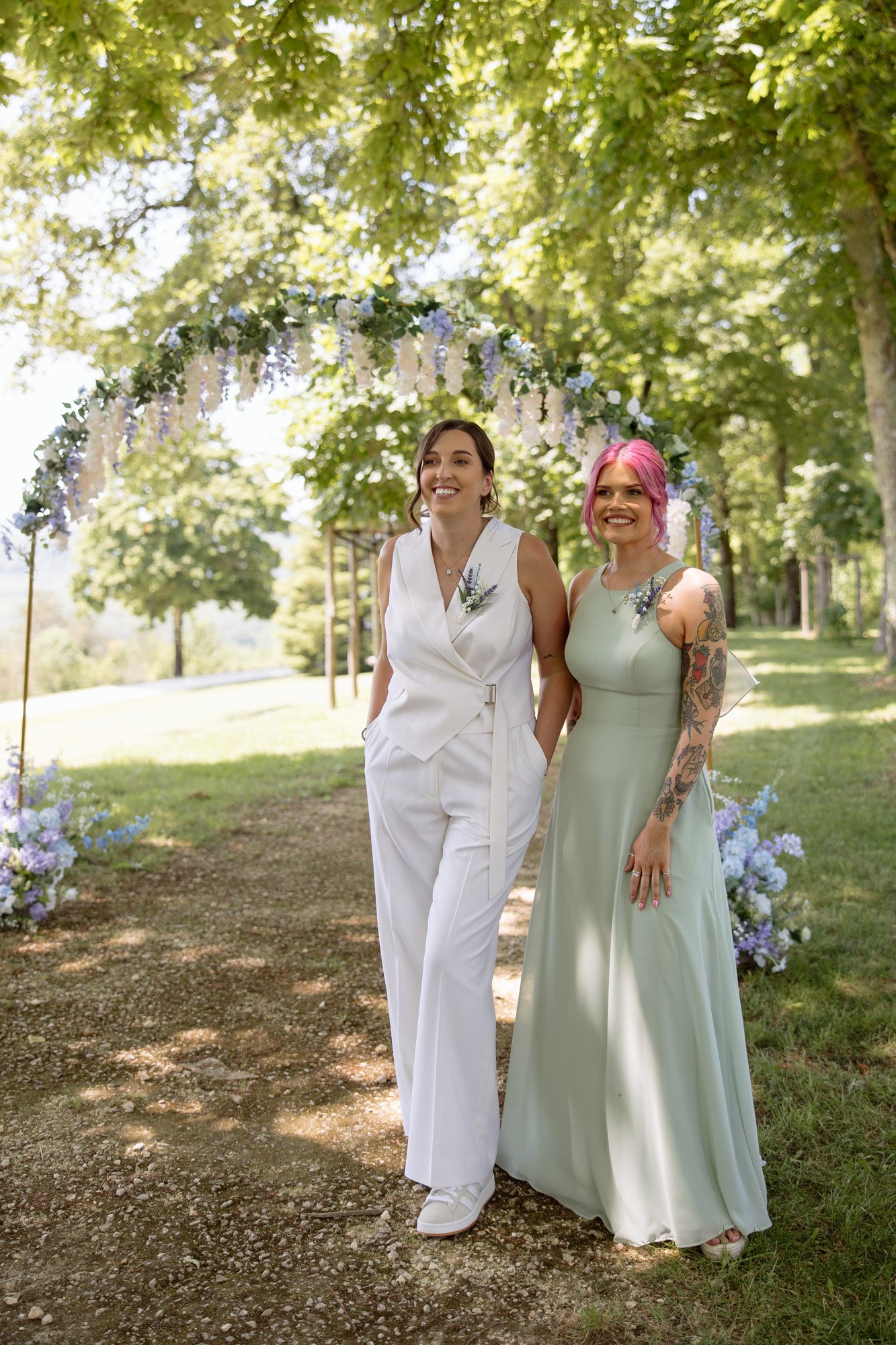 Two people stand under a floral arch outdoors; one wears a white suit and the other a light green dress. Both are smiling, surrounded by greenery and trees in the background.