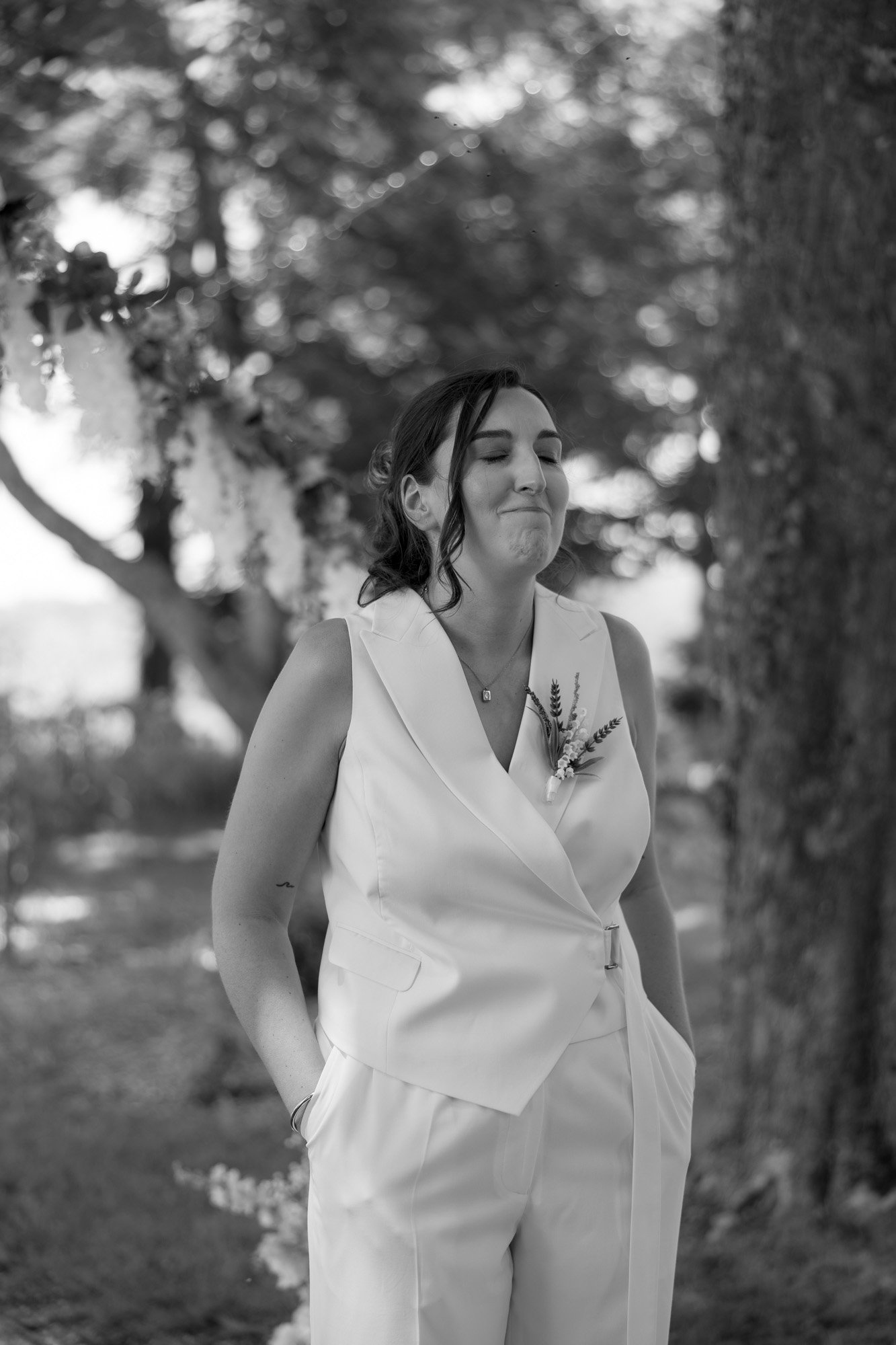 A person in a white suit stands outdoors with hands in pockets, eyes closed, and a neutral expression. Trees and foliage are visible in the background. Spring Dordogne Wedding Photographer