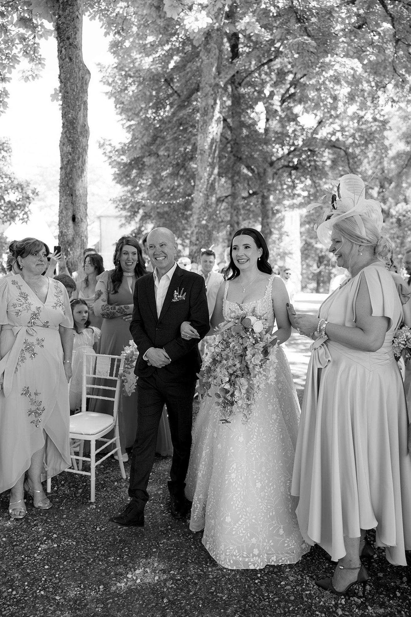 A bride in a white dress walks down an outdoor aisle on the arm of an older man, surrounded by guests dressed in formal attire, under a canopy of trees. Spring Dordogne Wedding Photographer