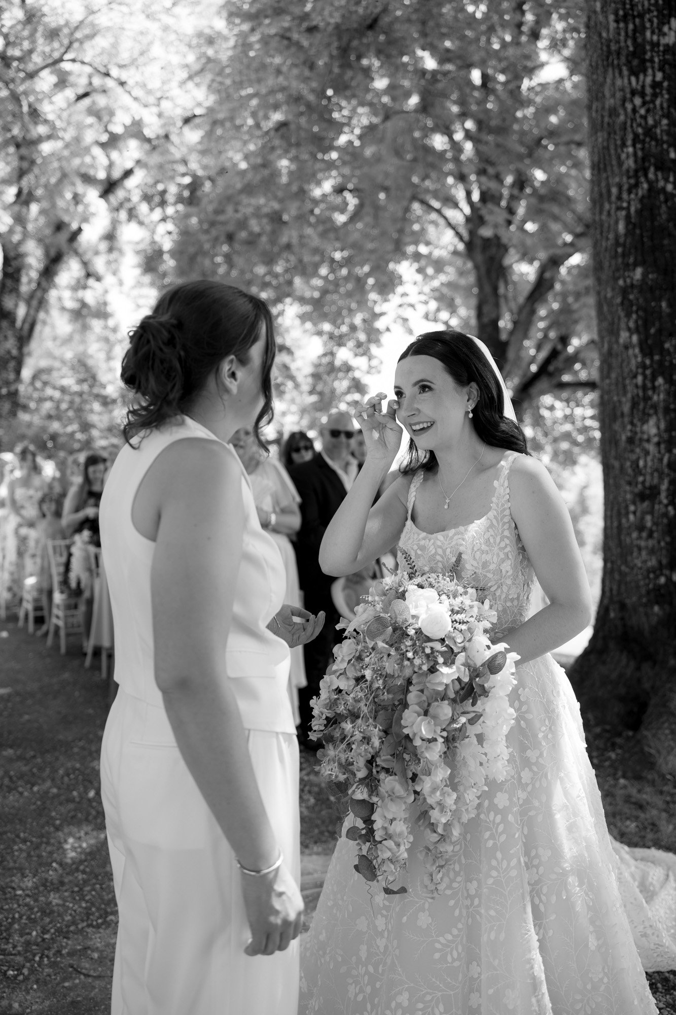 Two women stand outdoors at a wedding ceremony; one in a dress holding a bouquet, the other in a suit. Guests are seated in the background among trees. Spring Dordogne Wedding Photographer