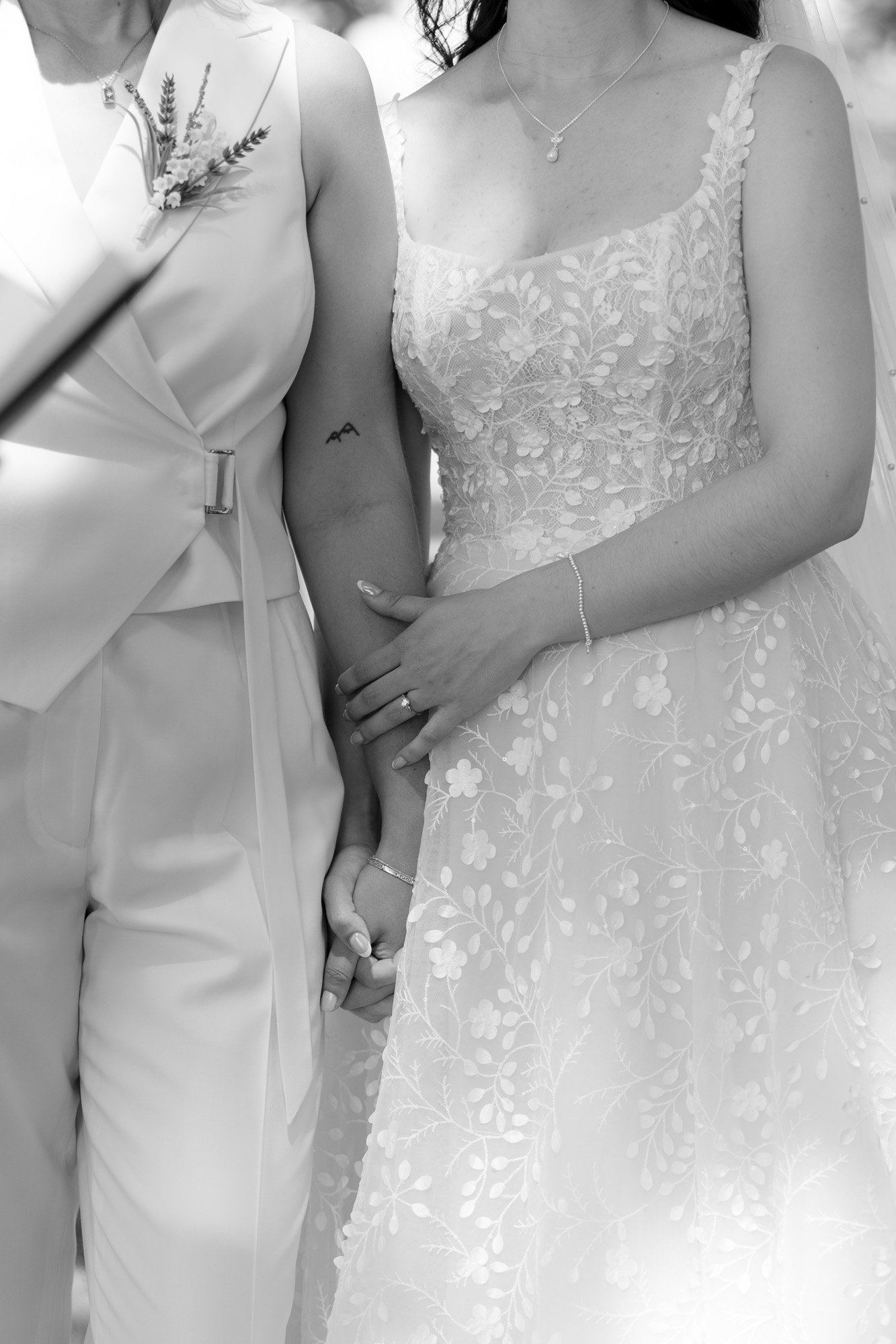 Two women in wedding attire, one in a suit and the other in a lace dress, hold hands and stand side by side.