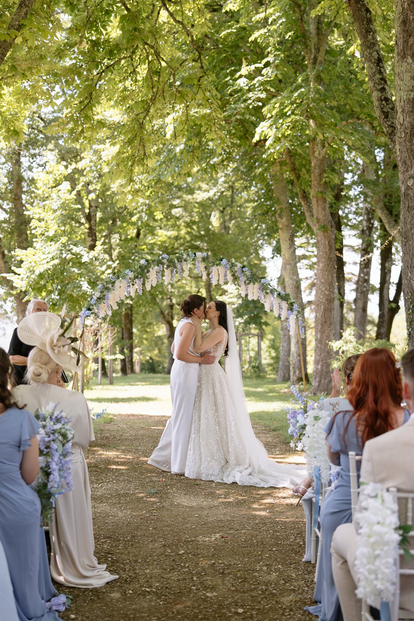 Two brides kiss under a floral arch during an outdoor wedding ceremony, surrounded by seated guests and tall green trees. Spring Dordogne Wedding Photographer