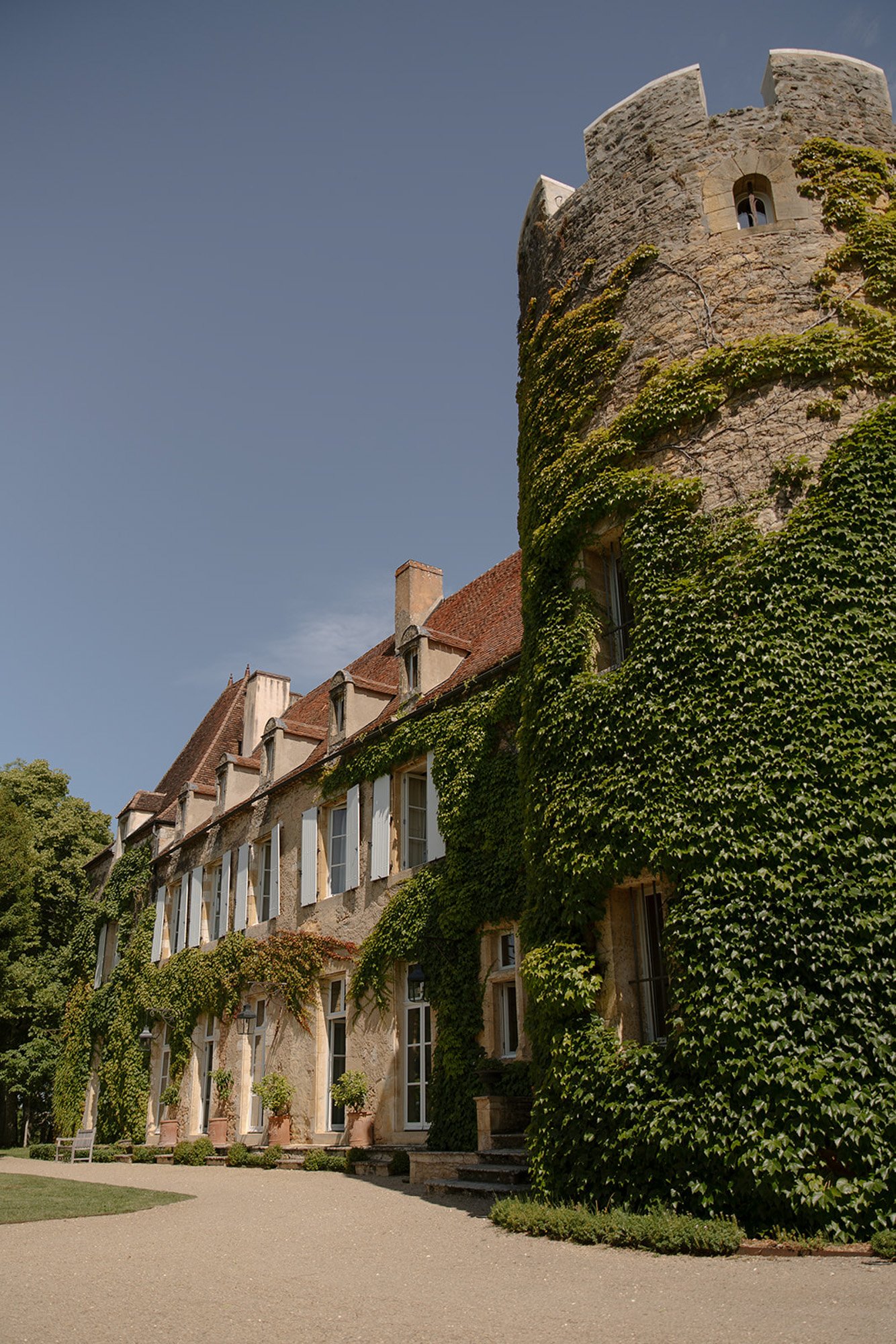 A stone building with a round tower is covered in green ivy, featuring white shutters and surrounded by greenery under a clear sky.