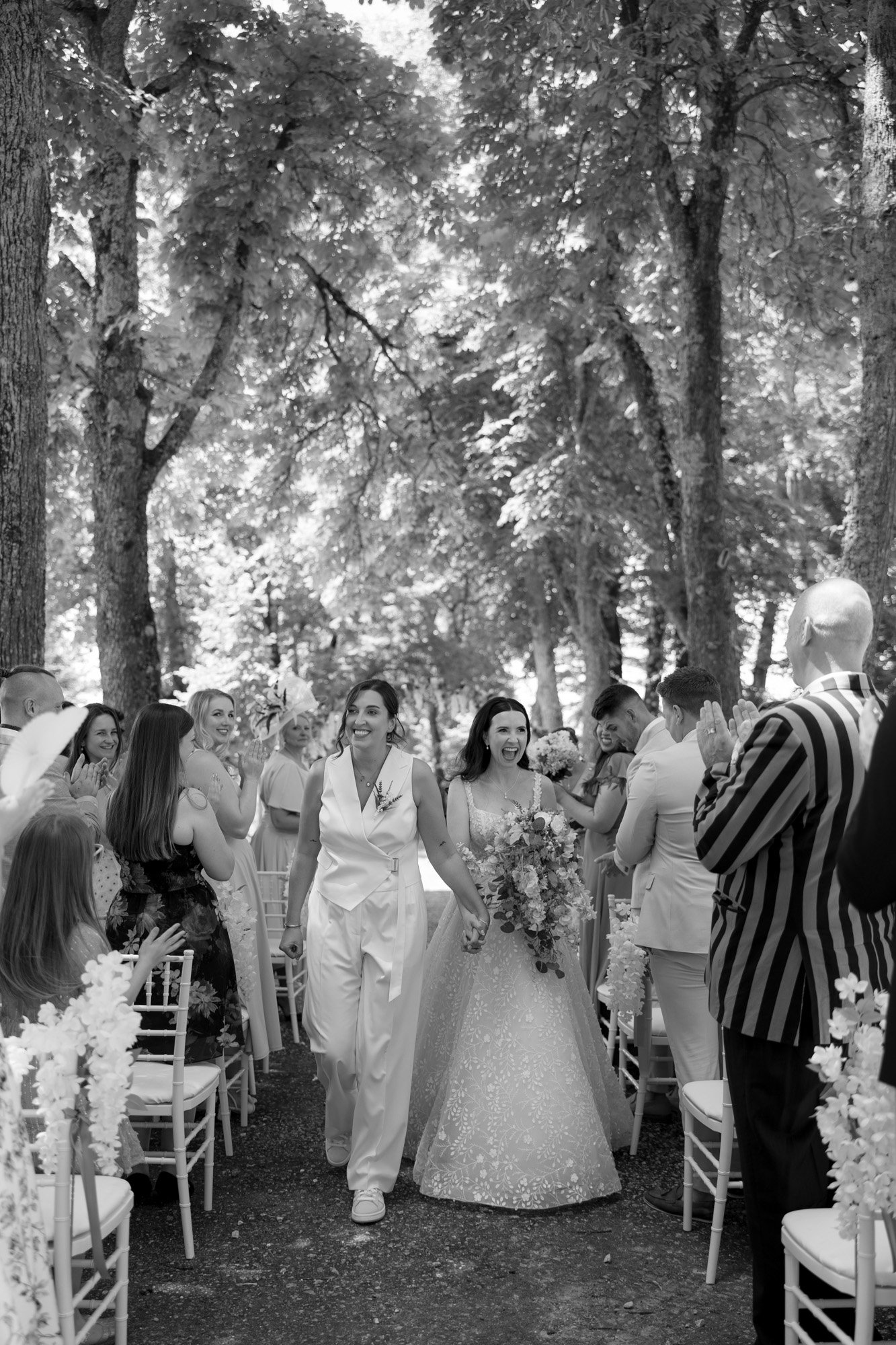 Two brides walk down the aisle together outdoors, surrounded by guests seated and standing among trees, clapping and celebrating their wedding. Spring Dordogne Wedding Photographer