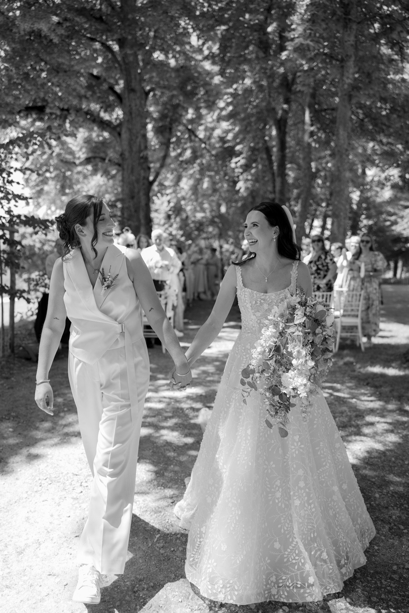 Two brides hold hands and smile at each other while walking outdoors during their wedding ceremony, surrounded by guests and trees. Spring Dordogne Wedding Photographer