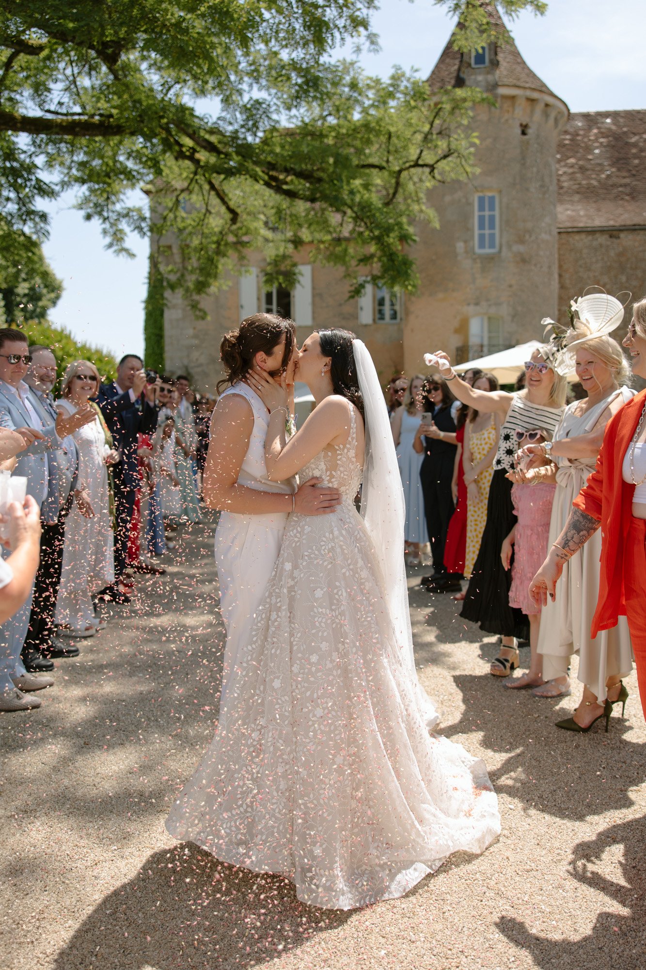 Two brides kiss outside at their wedding ceremony, surrounded by guests throwing confetti near a historic stone building.