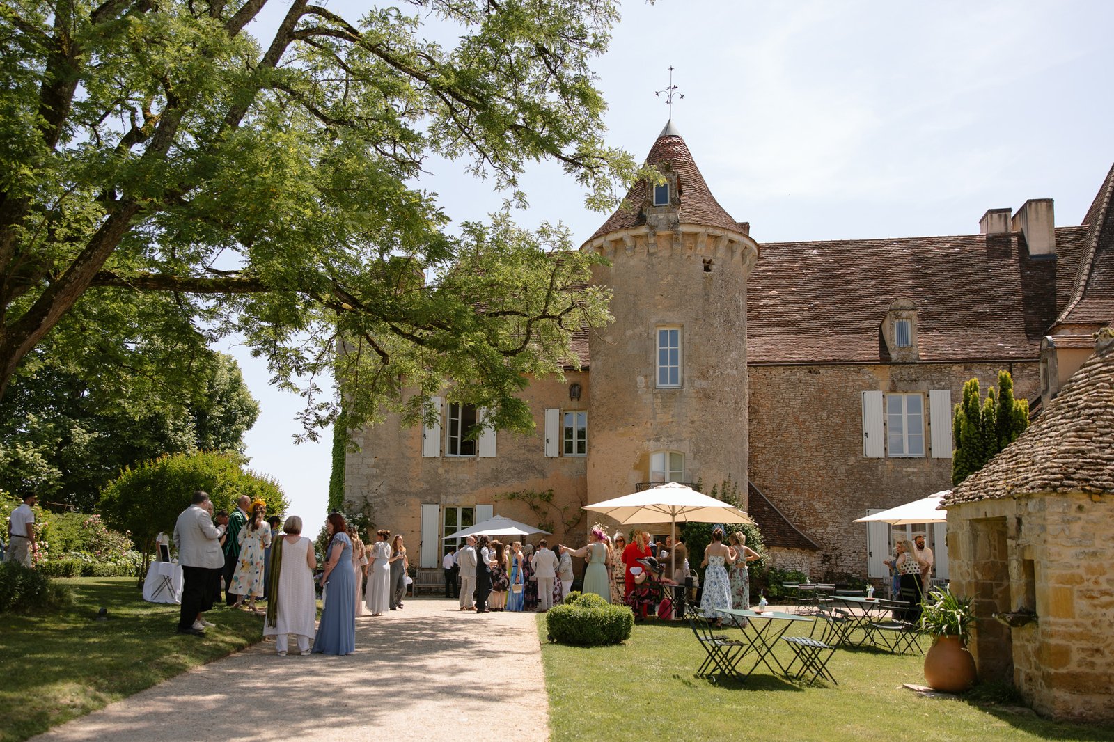 A group of people gathers outside a stone chateau with turrets on a sunny day, surrounded by trees, umbrellas, and garden furniture. Spring Dordogne Wedding Photographer