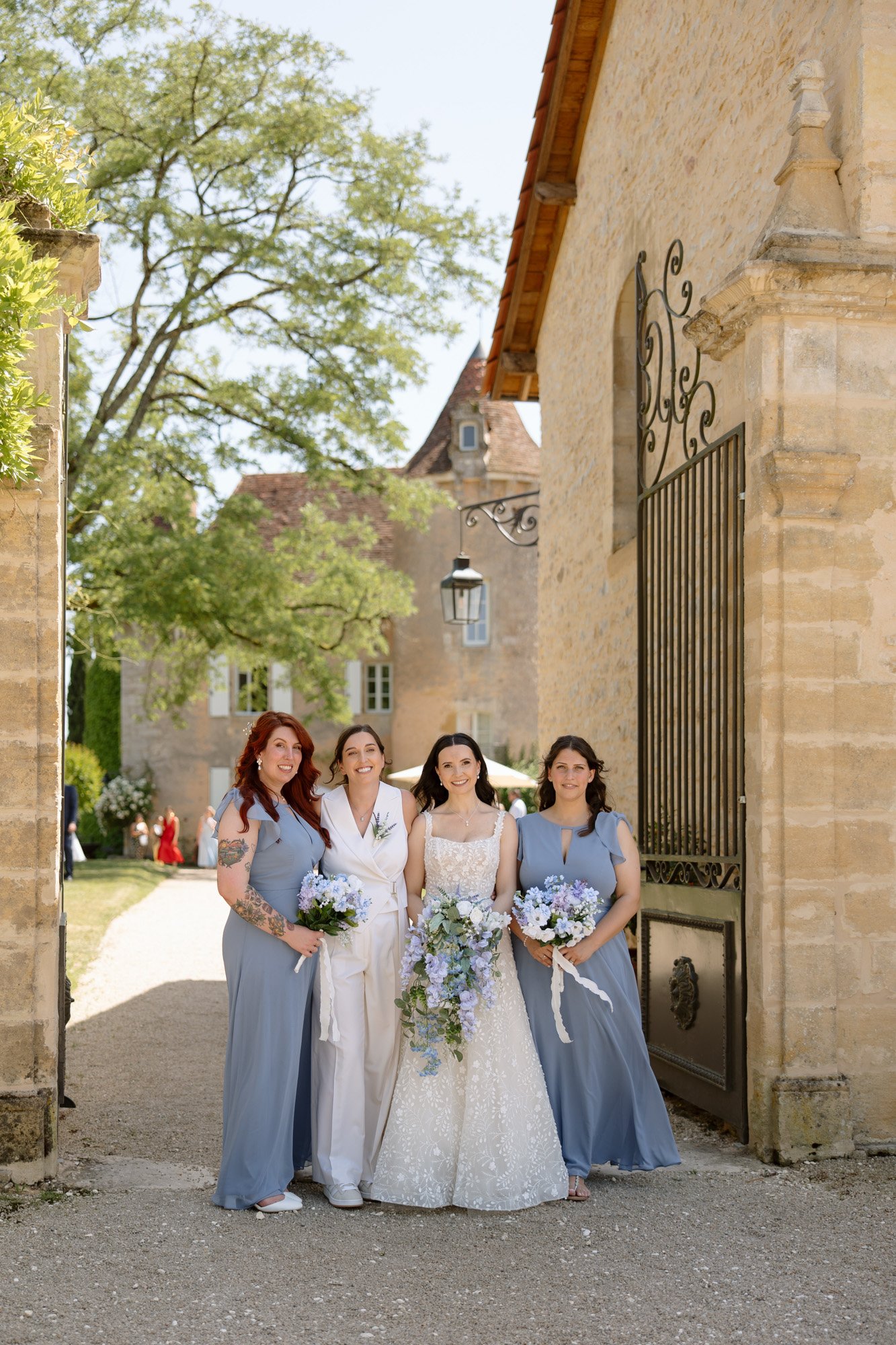 Four women stand outdoors in front of stone buildings; one wears a wedding dress, another a white suit, and two wear matching blue dresses, all holding bouquets. Spring Dordogne Wedding Photographer