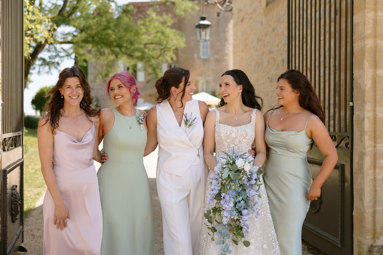 Five women stand together outdoors in formal attire, with one holding a bouquet of flowers. They are smiling and appear to be at a wedding or formal event.