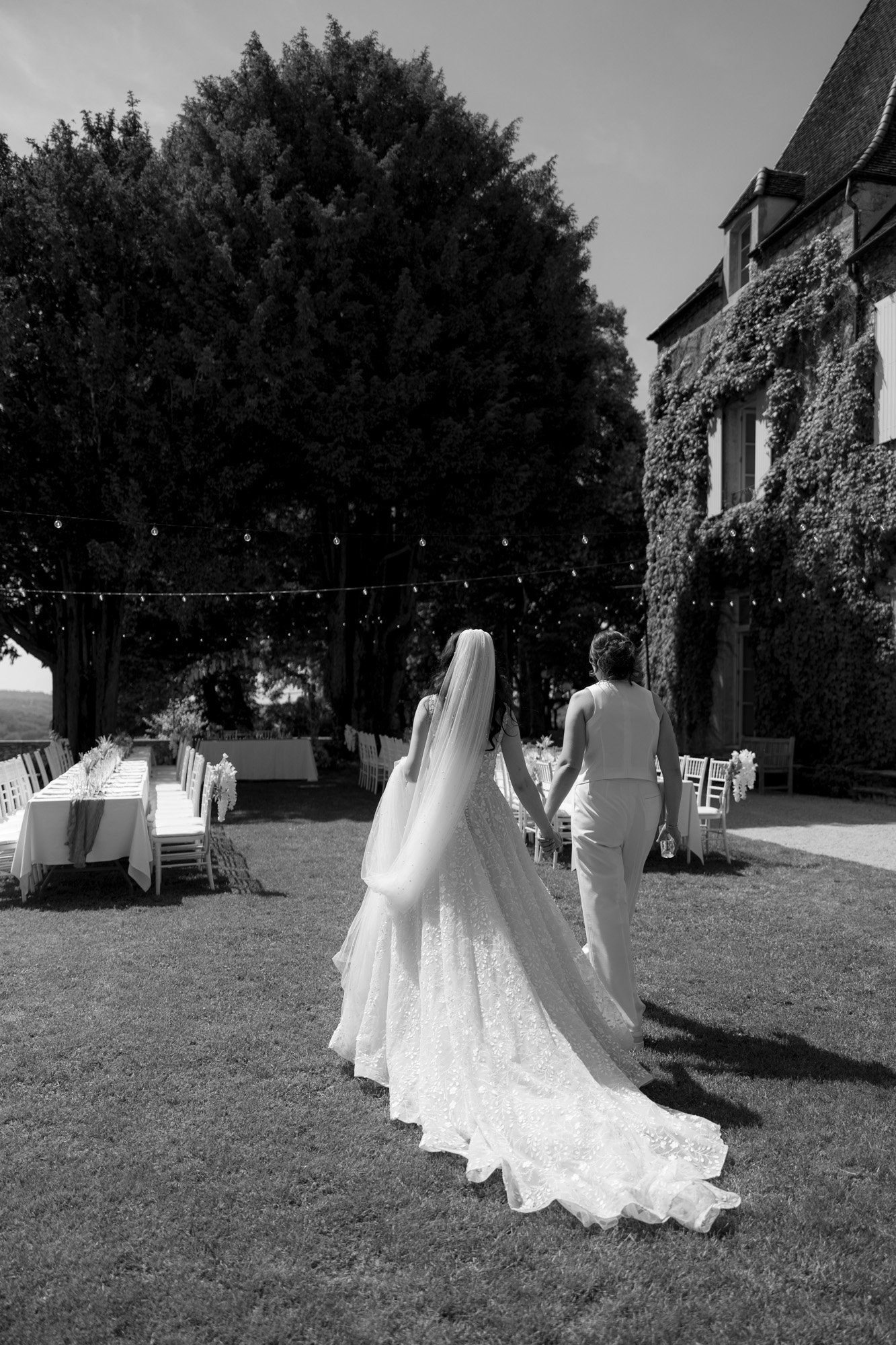 Two women, one in a wedding dress and the other in a suit, walk hand in hand on a grassy lawn near an ivy-covered building, with decorated tables and string lights nearby. Spring Dordogne Wedding Photographer