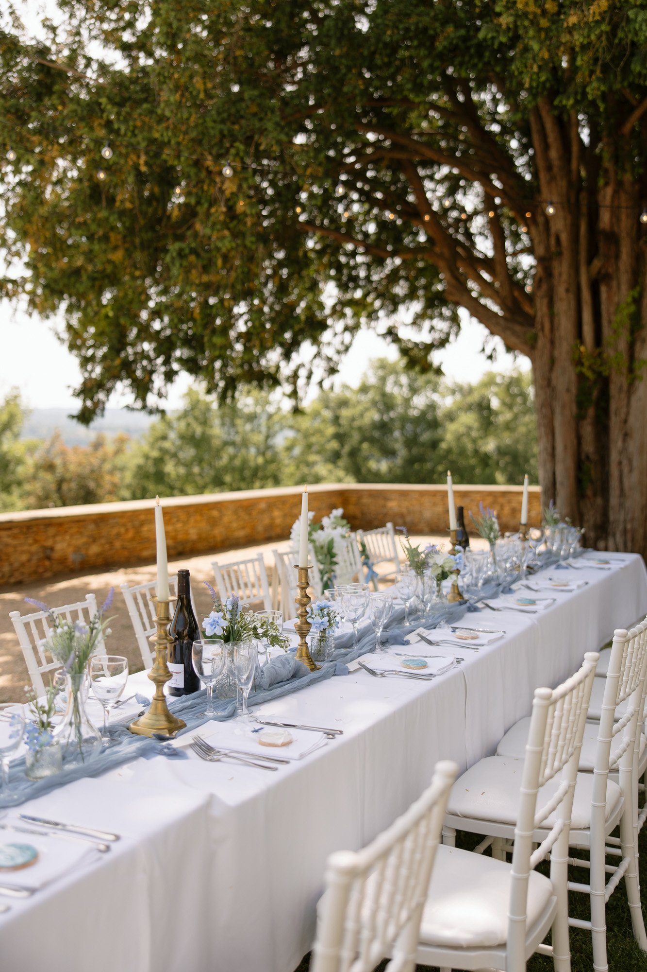 A long outdoor dining table with a white tablecloth is set with glassware, plates, candles, and floral centerpieces beside a large tree in a garden setting. Spring Dordogne Wedding Photographer