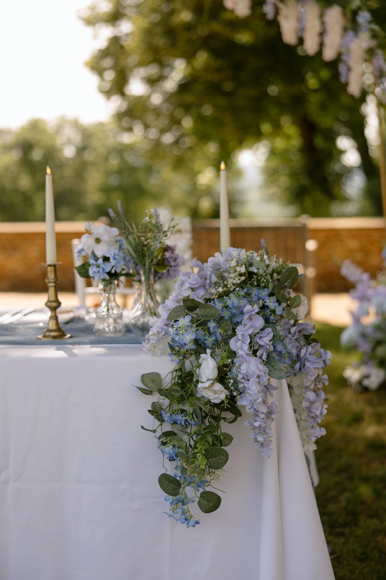 A white table with a floral arrangement of purple and white flowers, two lit candles, and small vases, set outdoors near a stone wall and trees.