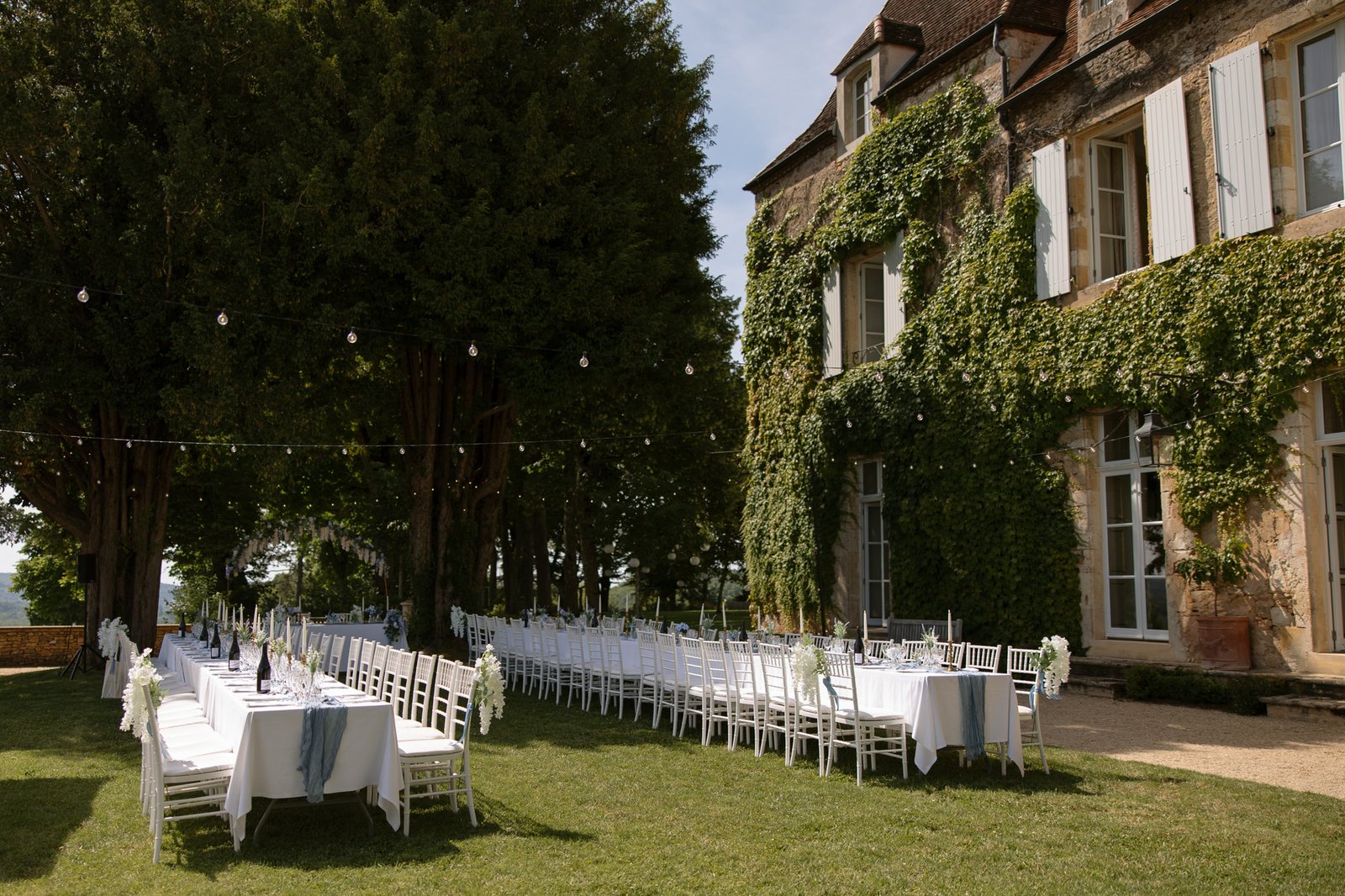 Long banquet tables with white chairs are set outdoors on a lawn beside an ivy-covered building, decorated with white linens and string lights overhead.