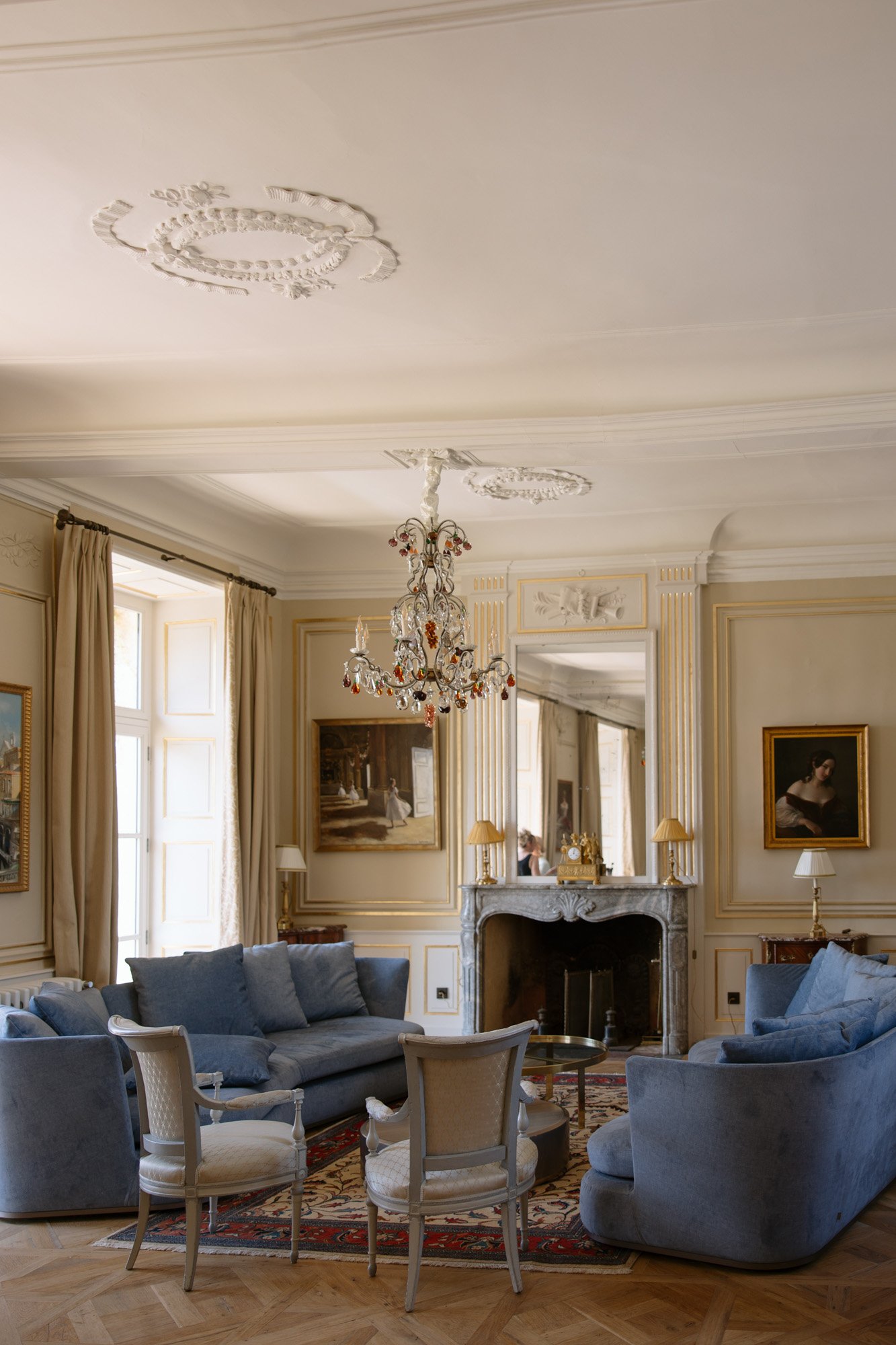 Elegant living room with ornate ceiling, chandelier, blue sofas, armchairs, fireplace, large mirror, paintings, and tall windows with beige curtains.