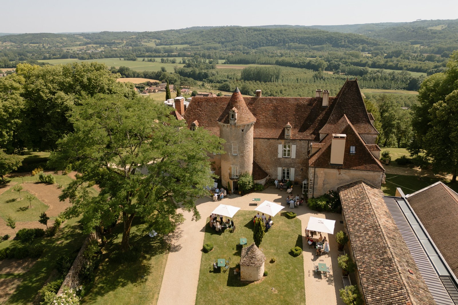 Aerial view of a historic stone chateau with a red-tiled roof, surrounded by trees, gardens, and people gathering under white umbrellas in the courtyard. Rolling hills in the background. Spring Dordogne Wedding Photographer