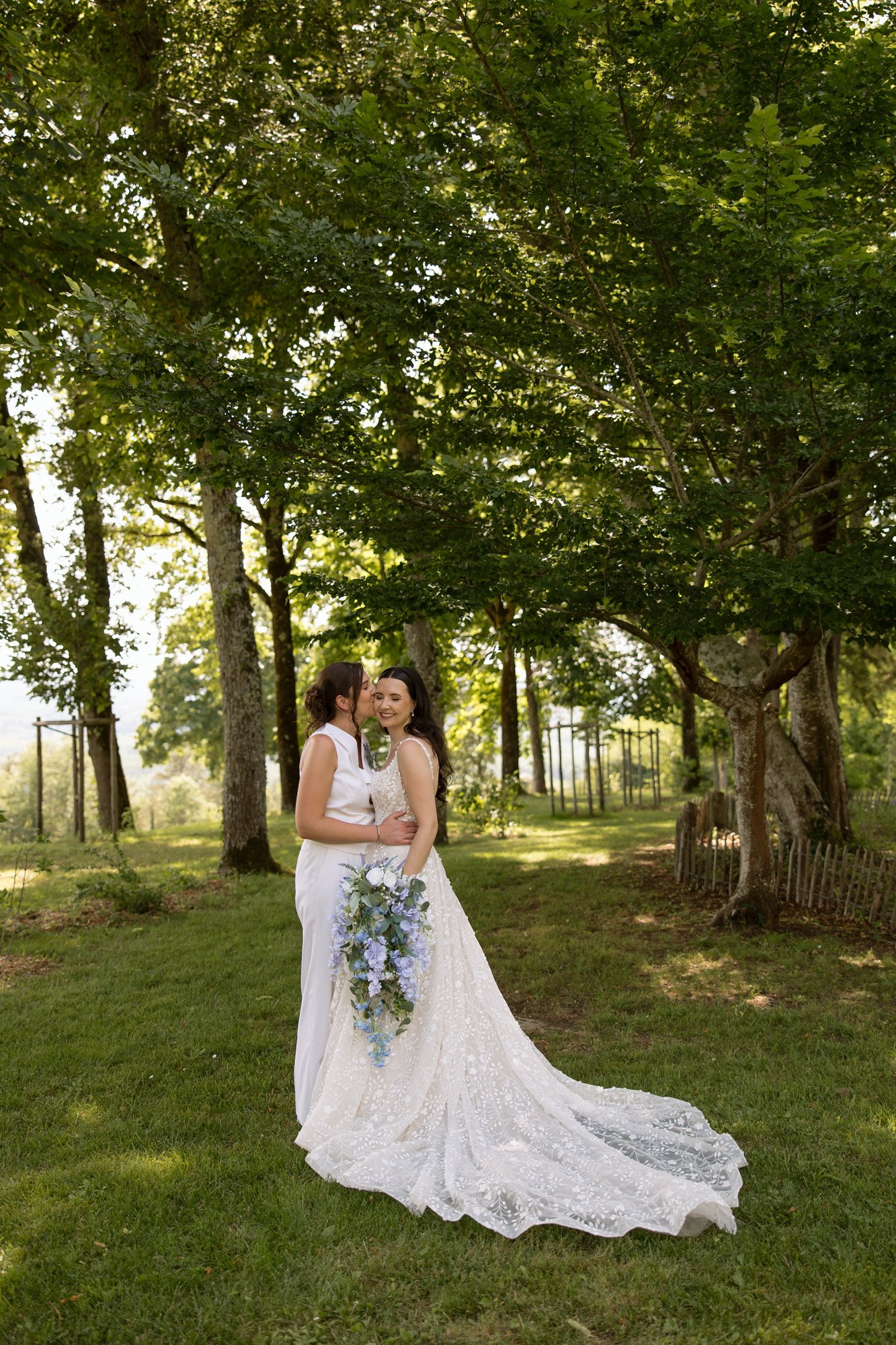 Two women in wedding attire stand together outdoors on grass, surrounded by trees. One holds a bouquet of blue and white flowers, and both are smiling. Spring Dordogne Wedding Photographer
