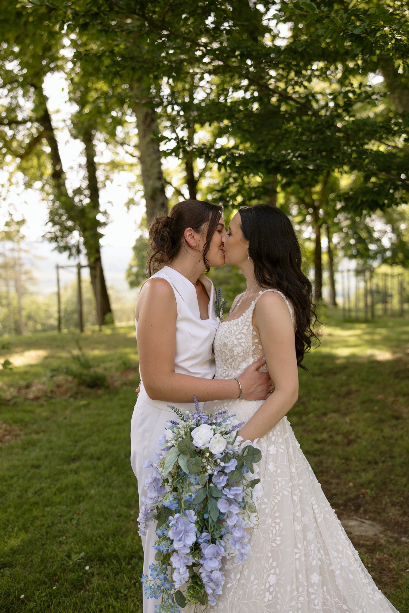 Two women in wedding attire share a kiss outdoors in a sunlit, wooded area. One wears a dress, the other a suit, and they hold a bouquet of purple and white flowers.