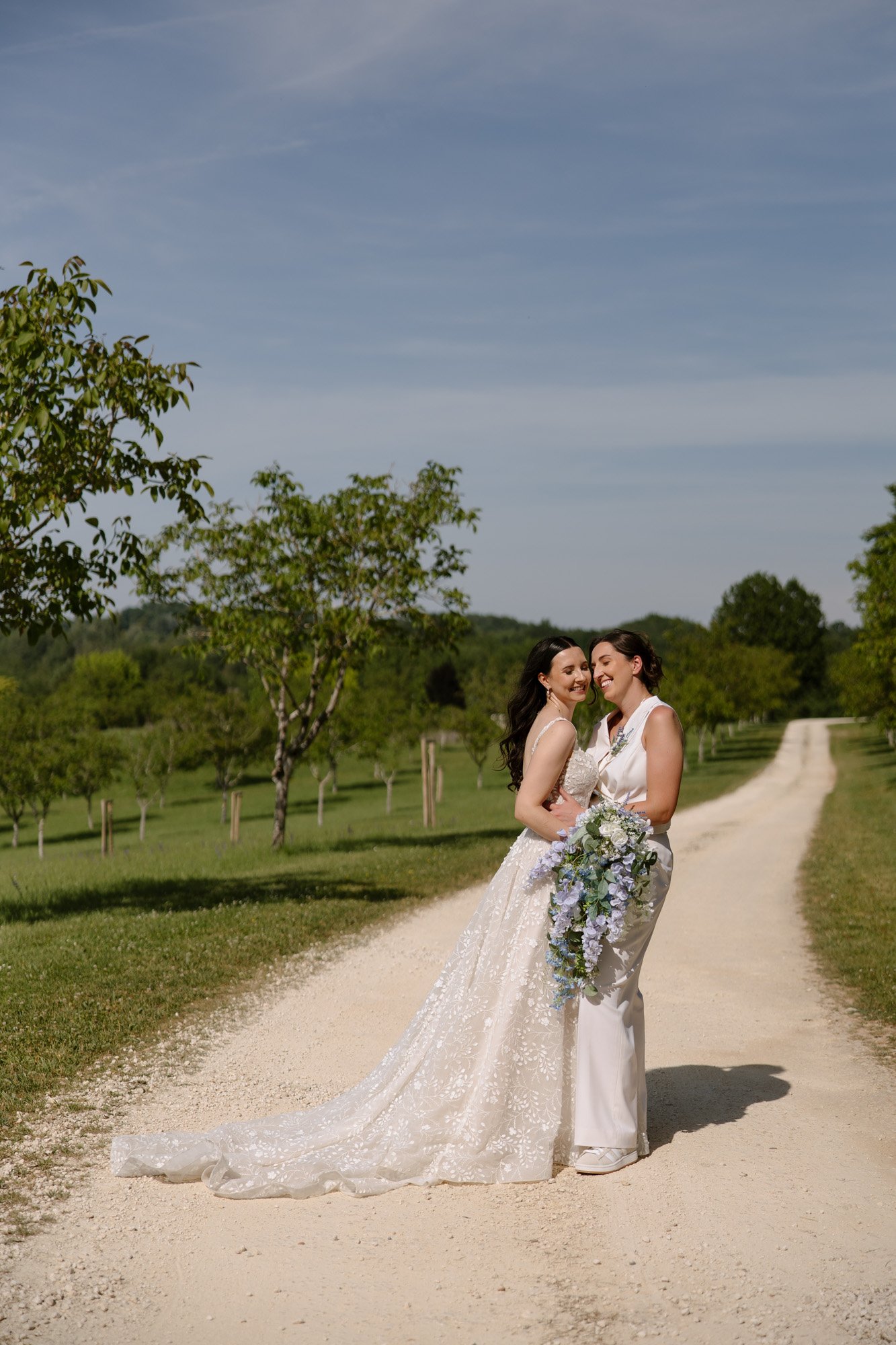 Two women in wedding attire stand together on a rural gravel path, one in a gown and the other in a suit, holding a bouquet, with greenery and trees in the background. Spring Dordogne Wedding Photographer