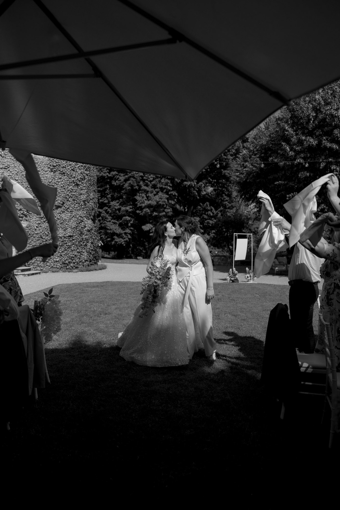 Two women in wedding dresses kiss outdoors under a large umbrella, while guests on either side wave white cloths in celebration. Spring Dordogne Wedding Photographer