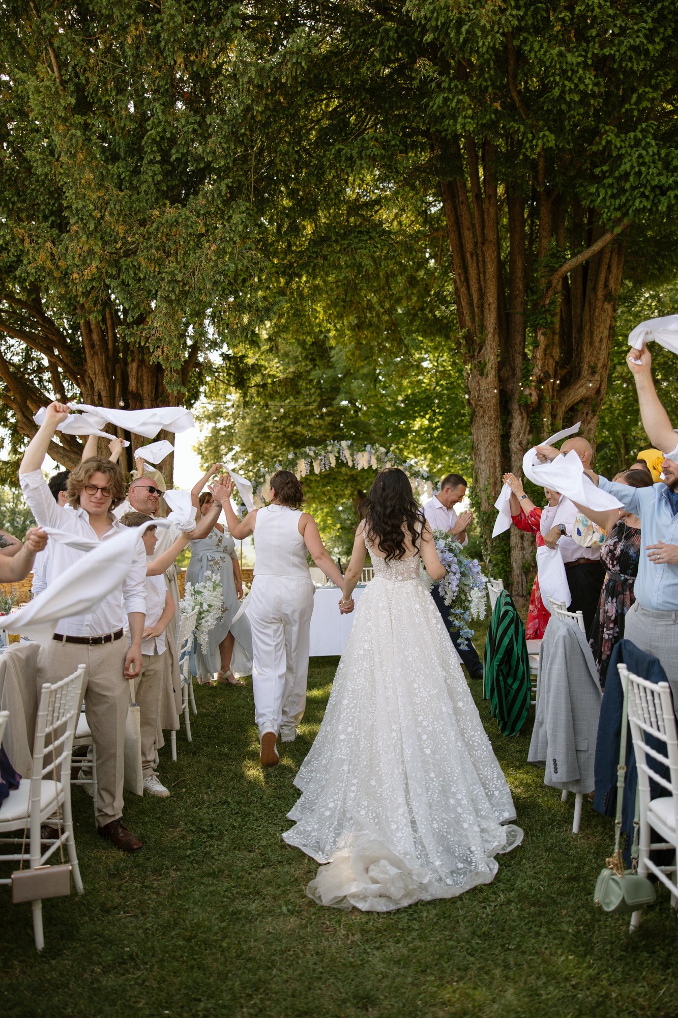 A bride and another person walk between outdoor tables while guests stand and wave white napkins in the air during a wedding celebration. Spring Dordogne Wedding Photographer