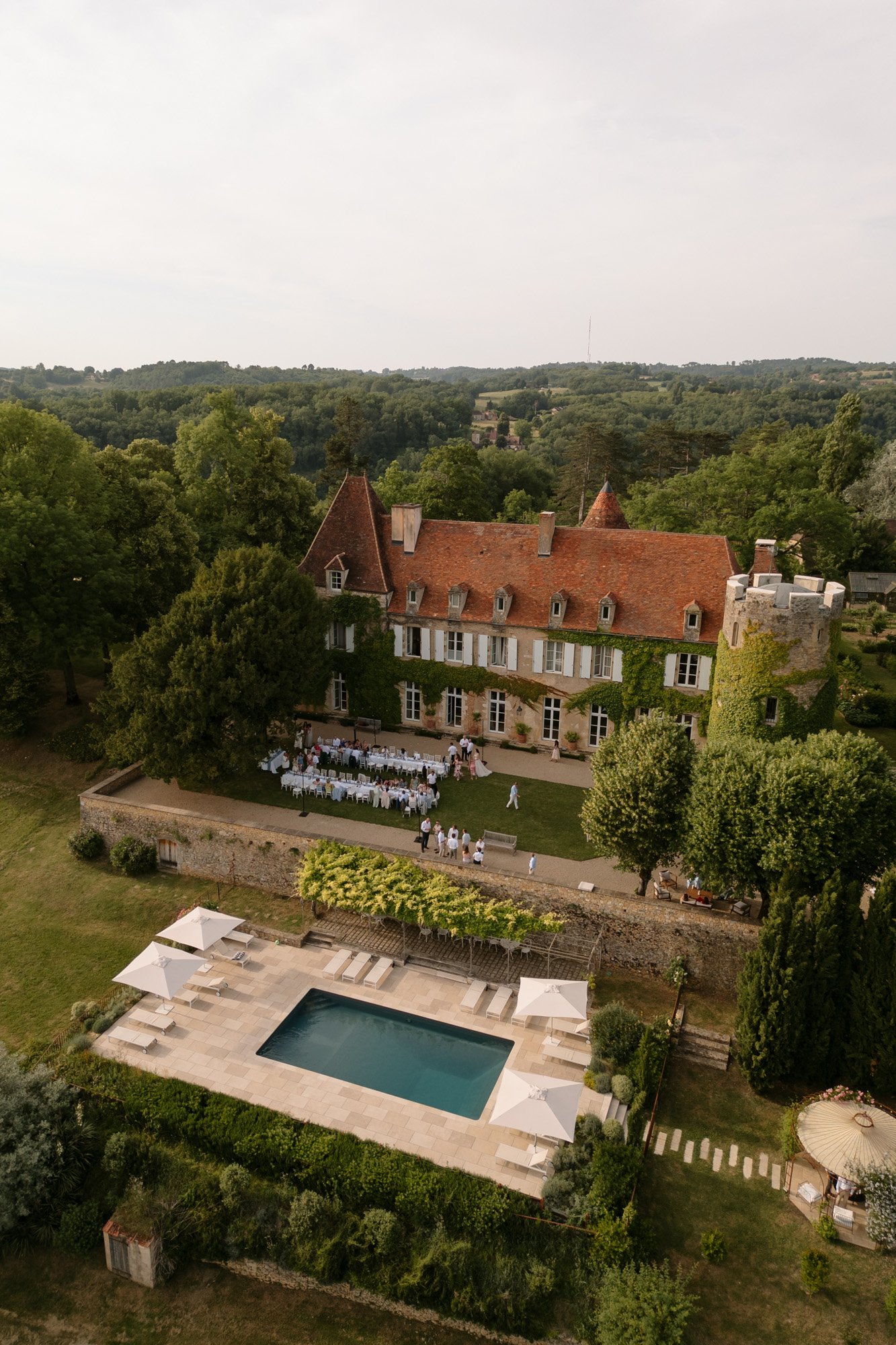 Aerial view of a large historic mansion with a red roof, surrounded by greenery, a group gathered on the patio, and a swimming pool with poolside umbrellas in the foreground.
