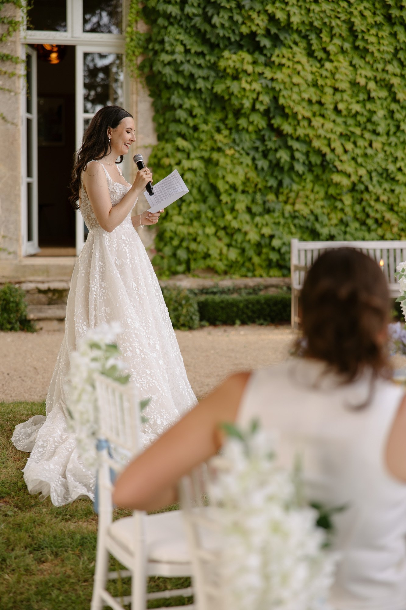 A bride in a white dress stands outdoors, holding a microphone and reading from a paper, while a seated guest looks on. Spring Dordogne Wedding Photographer