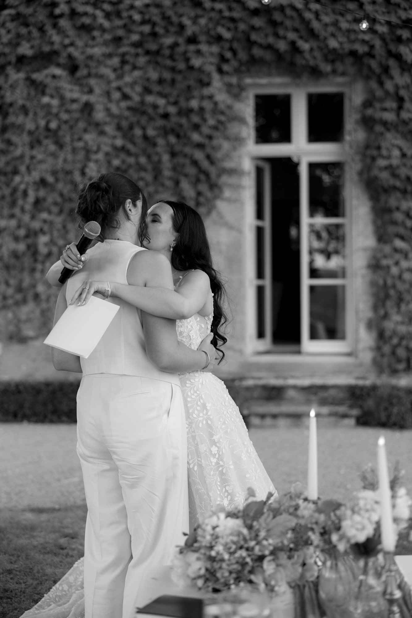 Two people embrace during an outdoor event; one holds a microphone and a piece of paper. A decorated table and building covered in ivy are visible in the background.