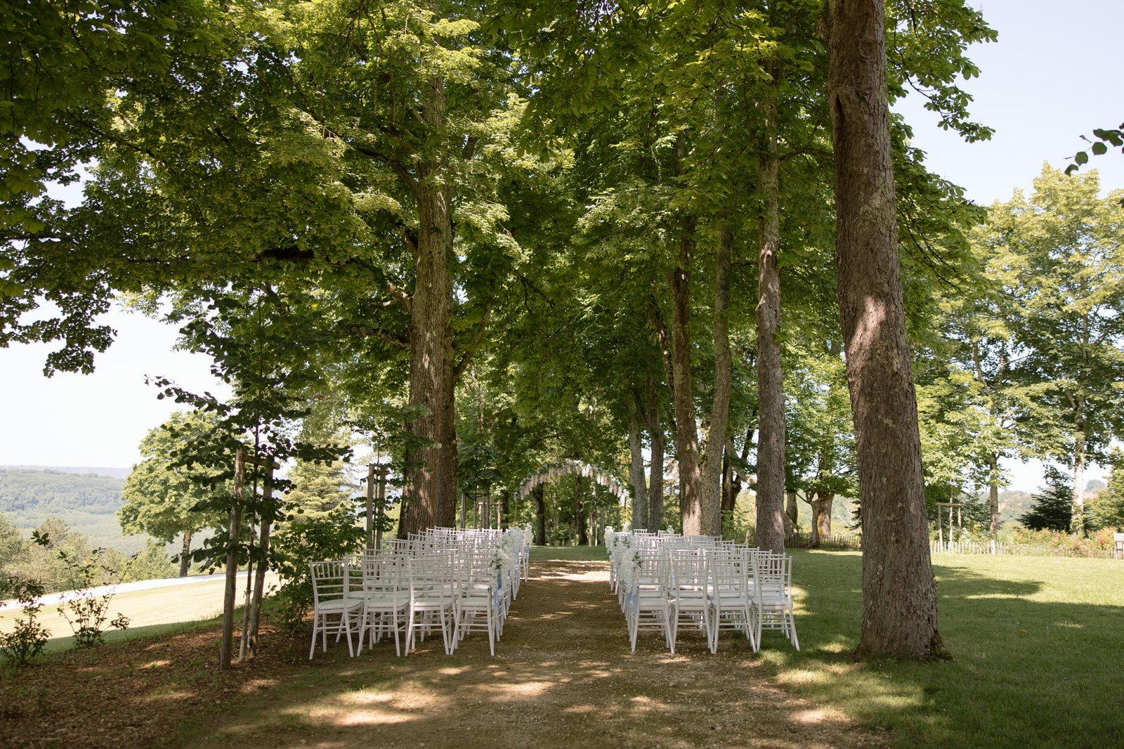 Rows of white chairs are set up along a tree-lined outdoor aisle on a dirt path, with green grass and distant landscape visible in the background.