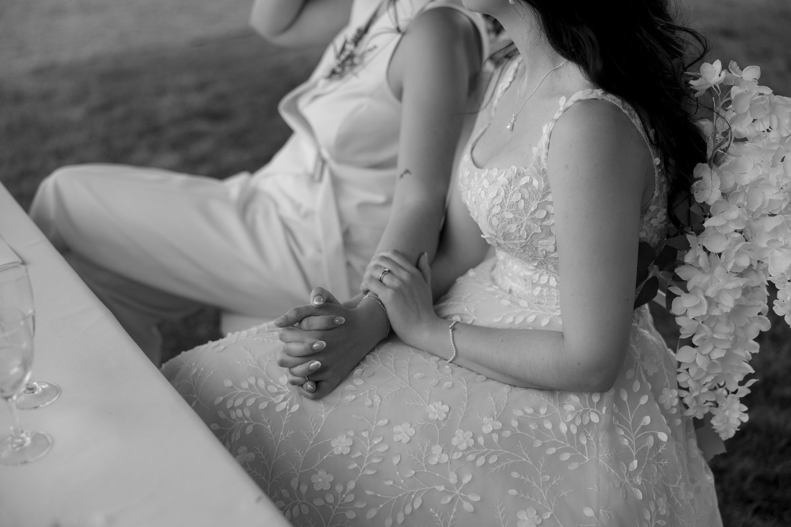 Two people sit closely together, holding hands. One wears a floral lace dress with a bouquet of white flowers, the other wears a suit. The image is in black and white.