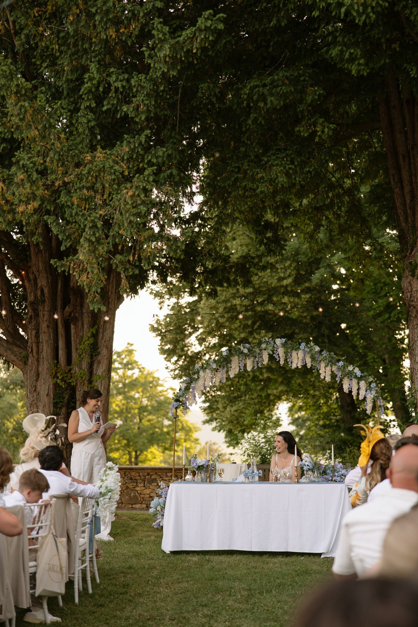 A person stands to deliver a speech outdoors at a wedding reception. The bride sits at a decorated table under a floral arch, surrounded by guests and trees. Spring Dordogne Wedding Photographer