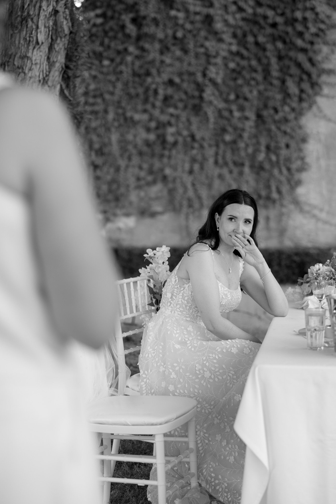 A woman in a light, patterned dress sits at an outdoor table, resting her hand on her mouth and looking ahead. Another person stands in the foreground, partially out of focus. Spring Dordogne Wedding Photographer