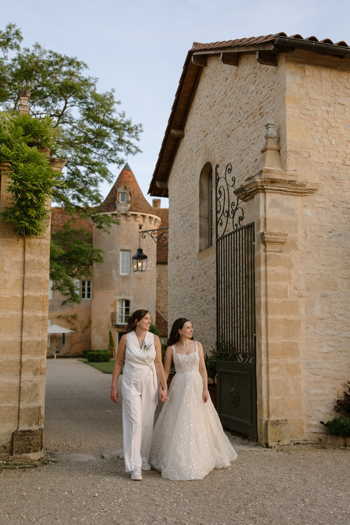 Two women, one in a white jumpsuit and the other in a wedding dress, walk hand in hand outside a stone building with an open wrought iron gate. Spring Dordogne Wedding Photographer