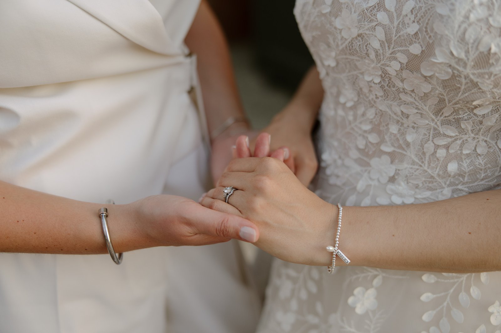 Two people stand close, holding hands. One wears a white lace dress and a pearl bracelet; the other wears a white suit and a silver bracelet. Both have manicured nails and rings. Spring Dordogne Wedding Photographer