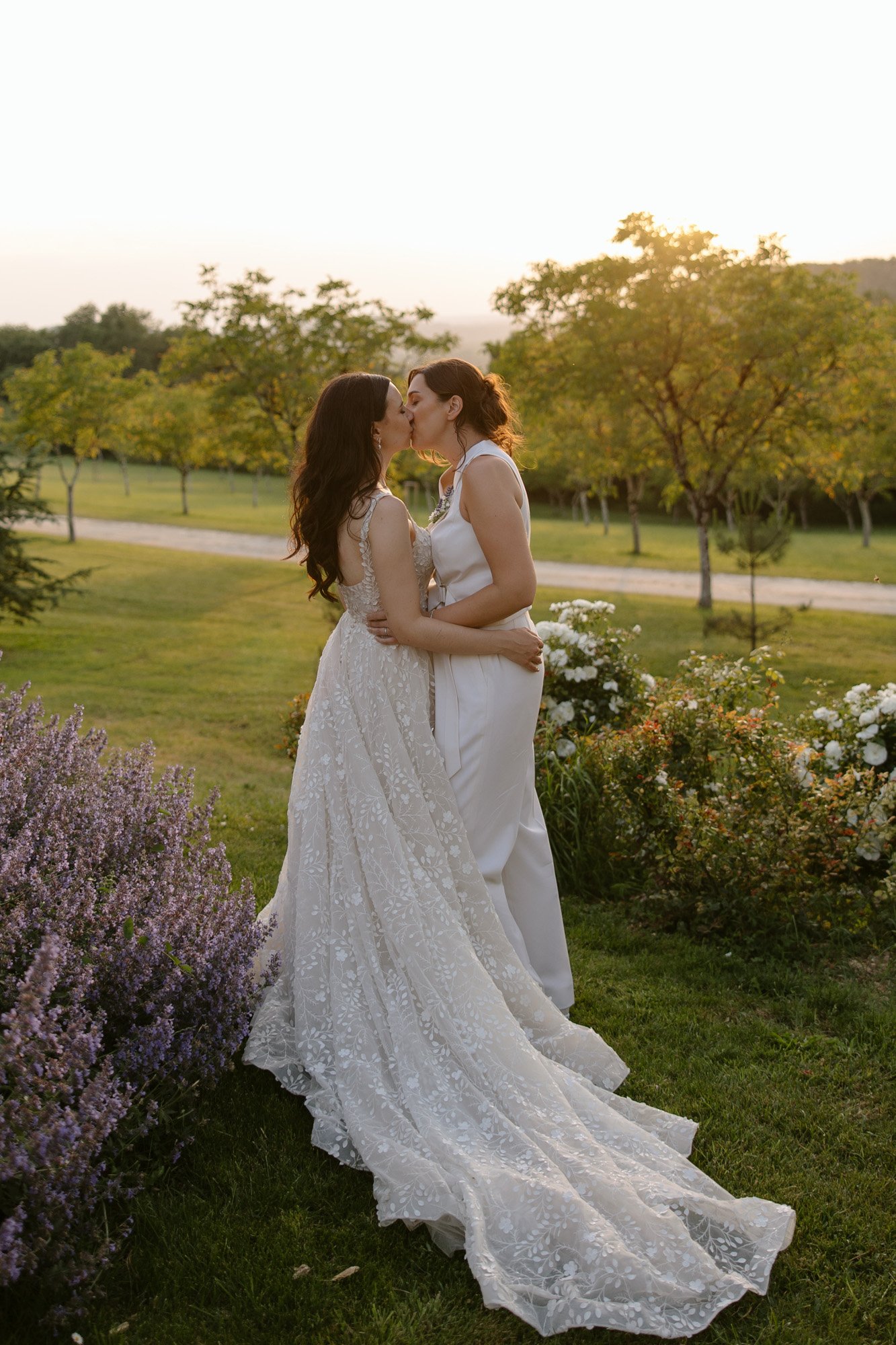 Two women in wedding attire stand outdoors at sunset, embracing and sharing a kiss in a garden with green grass and blooming flowers. Spring Dordogne Wedding Photographer