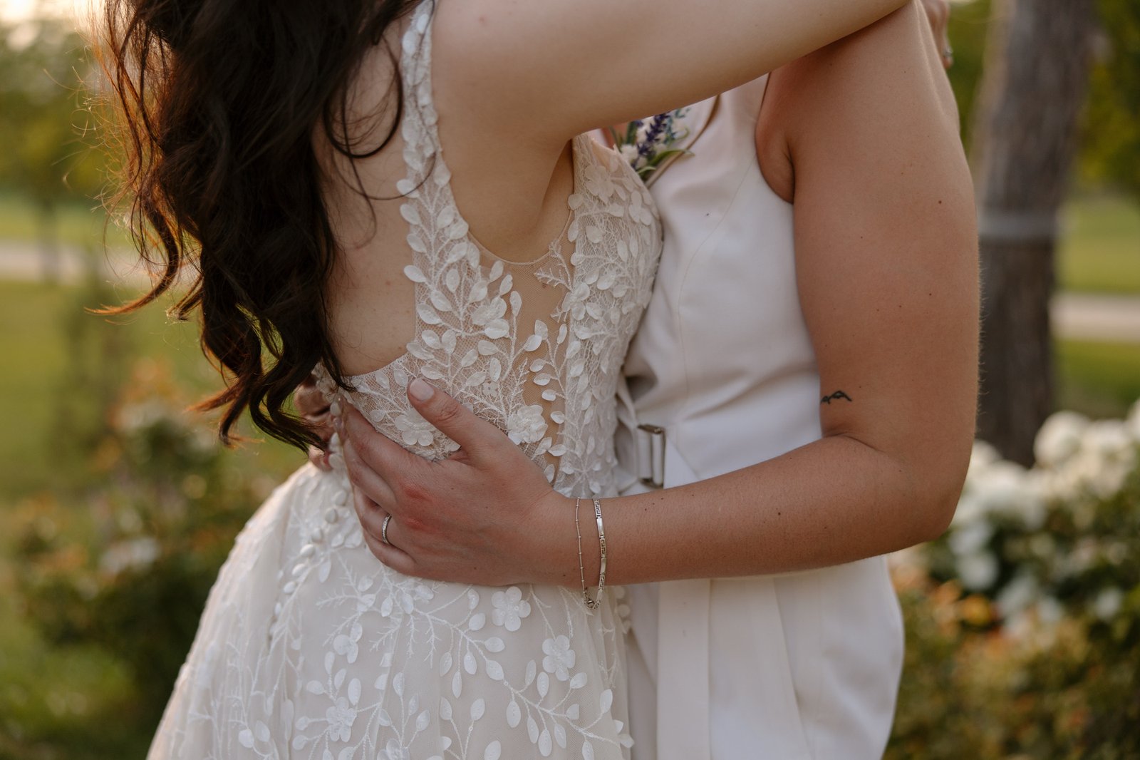 Two people in wedding attire embrace outdoors. One wears a sleeveless lace dress; the other wears a white sleeveless outfit. Their arms are wrapped around each other.