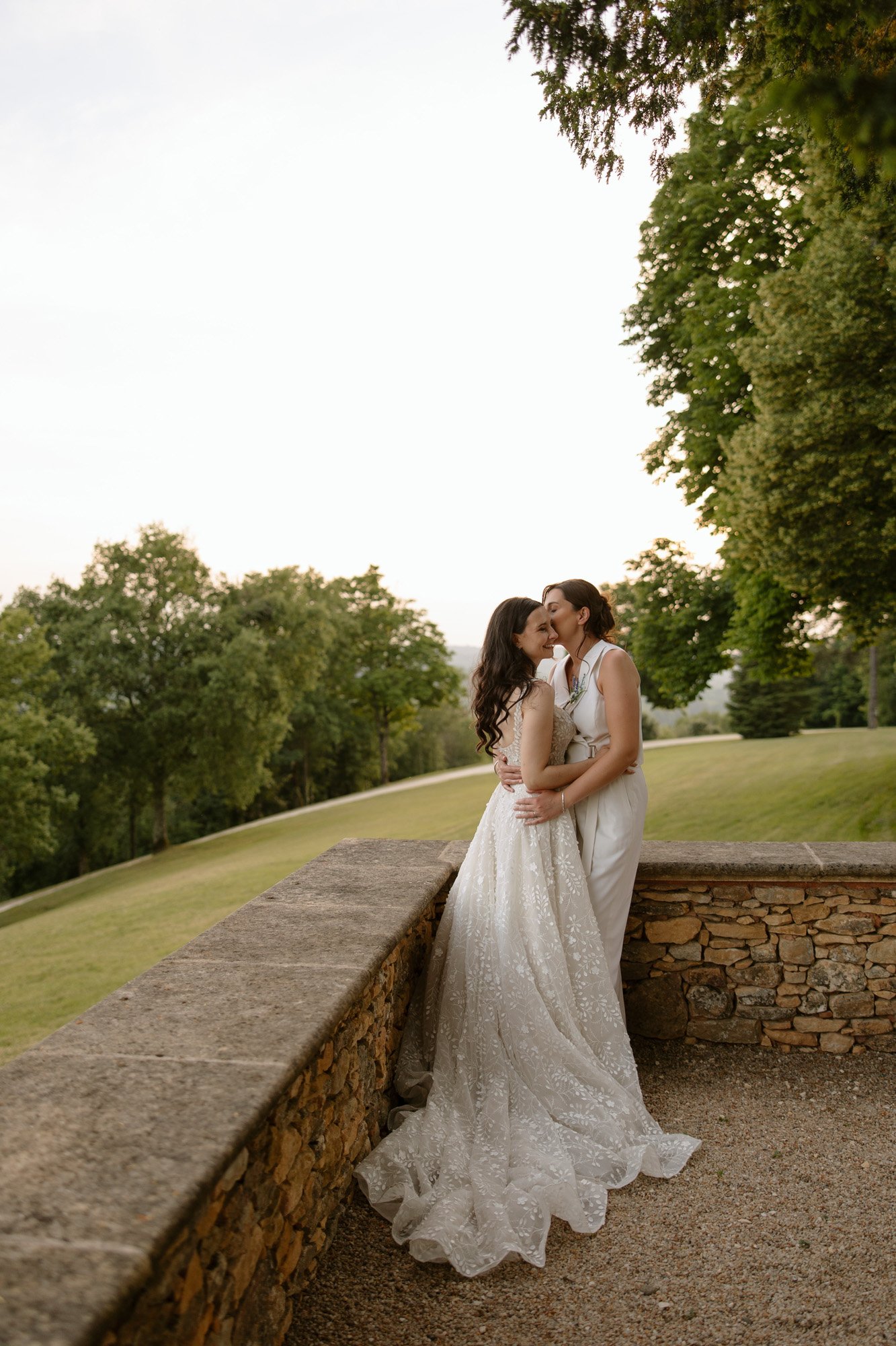 Two women in white dresses embrace and kiss outdoors by a stone wall, with trees and a grassy landscape in the background. Spring Dordogne Wedding Photographer