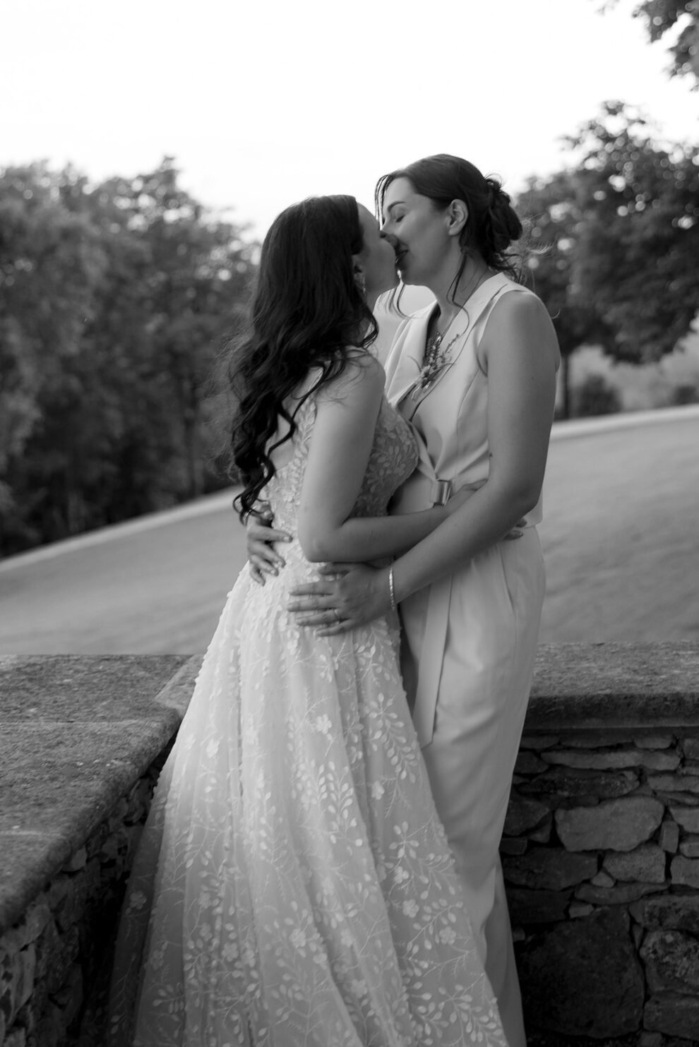 Two women in formal attire embrace and kiss outdoors near a stone wall, with trees and a pathway visible in the background.