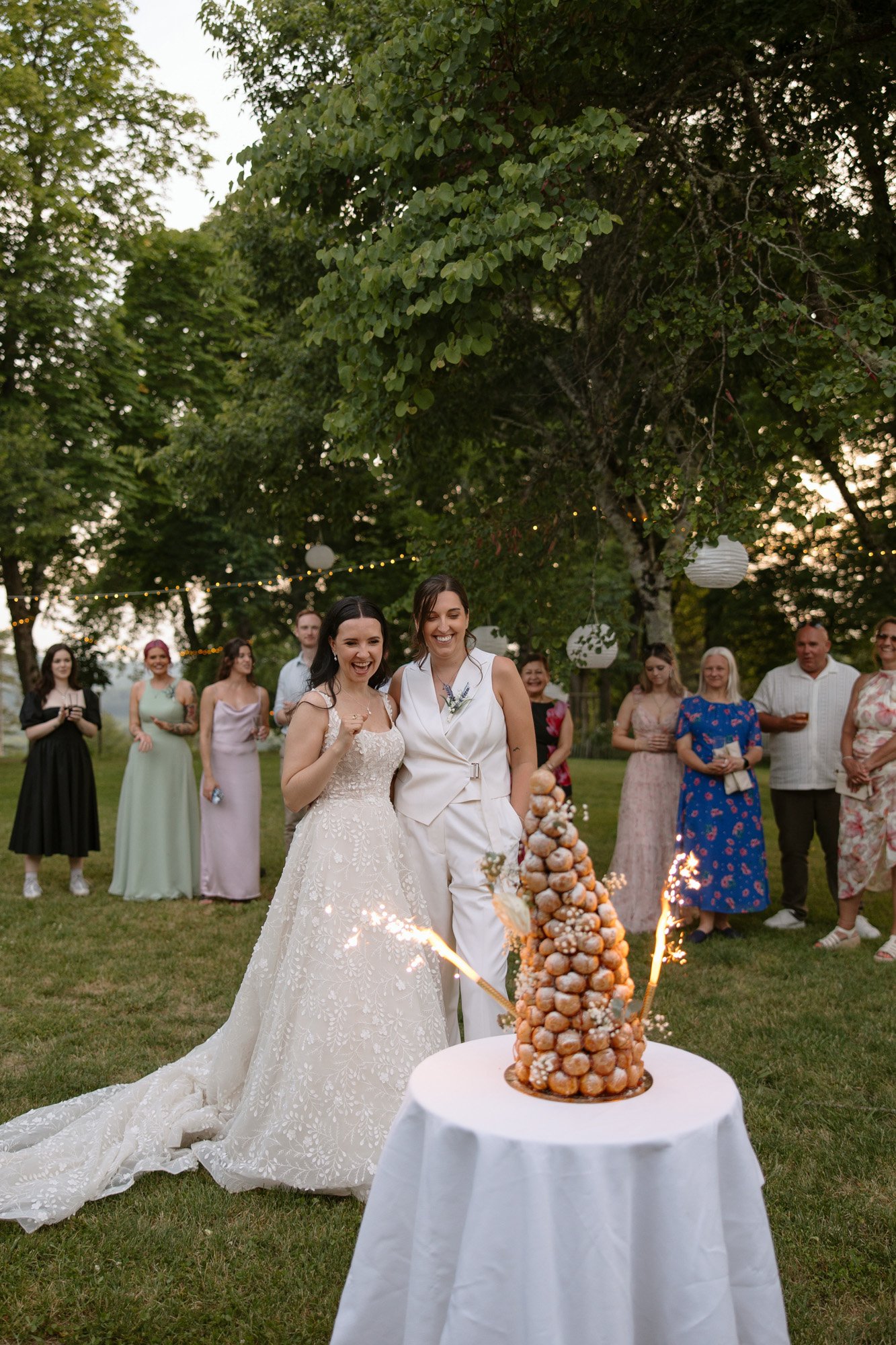 Two brides stand smiling in front of a croquembouche with sparklers, surrounded by guests outdoors at a wedding celebration. Spring Dordogne Wedding Photographer