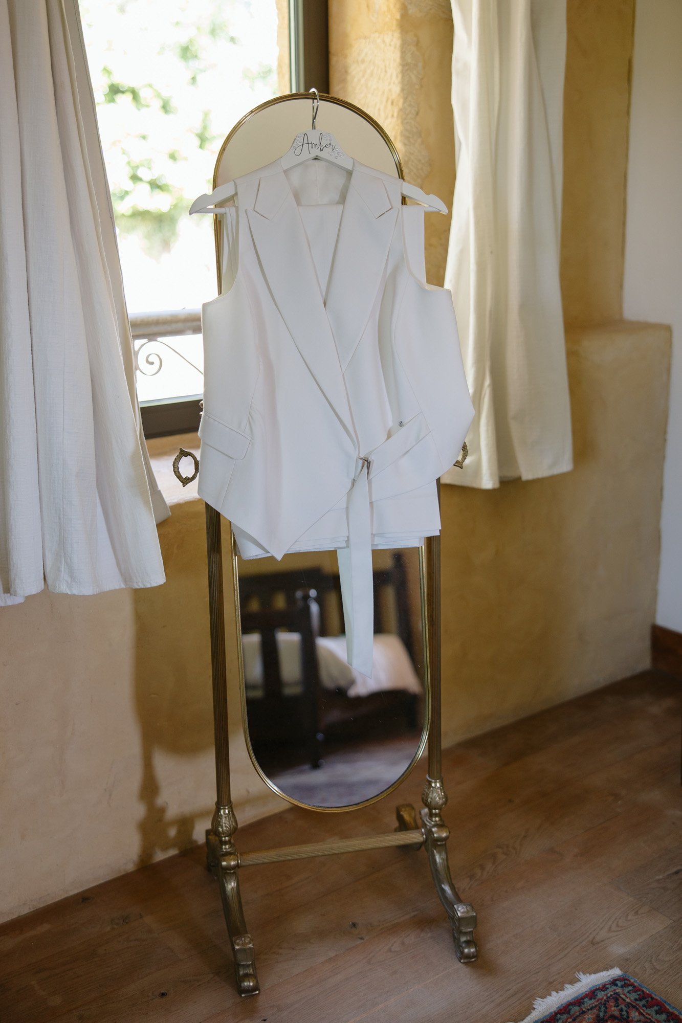 A white sleeveless blazer with a belt hangs on a hanger in front of a standing mirror near a window with curtains.