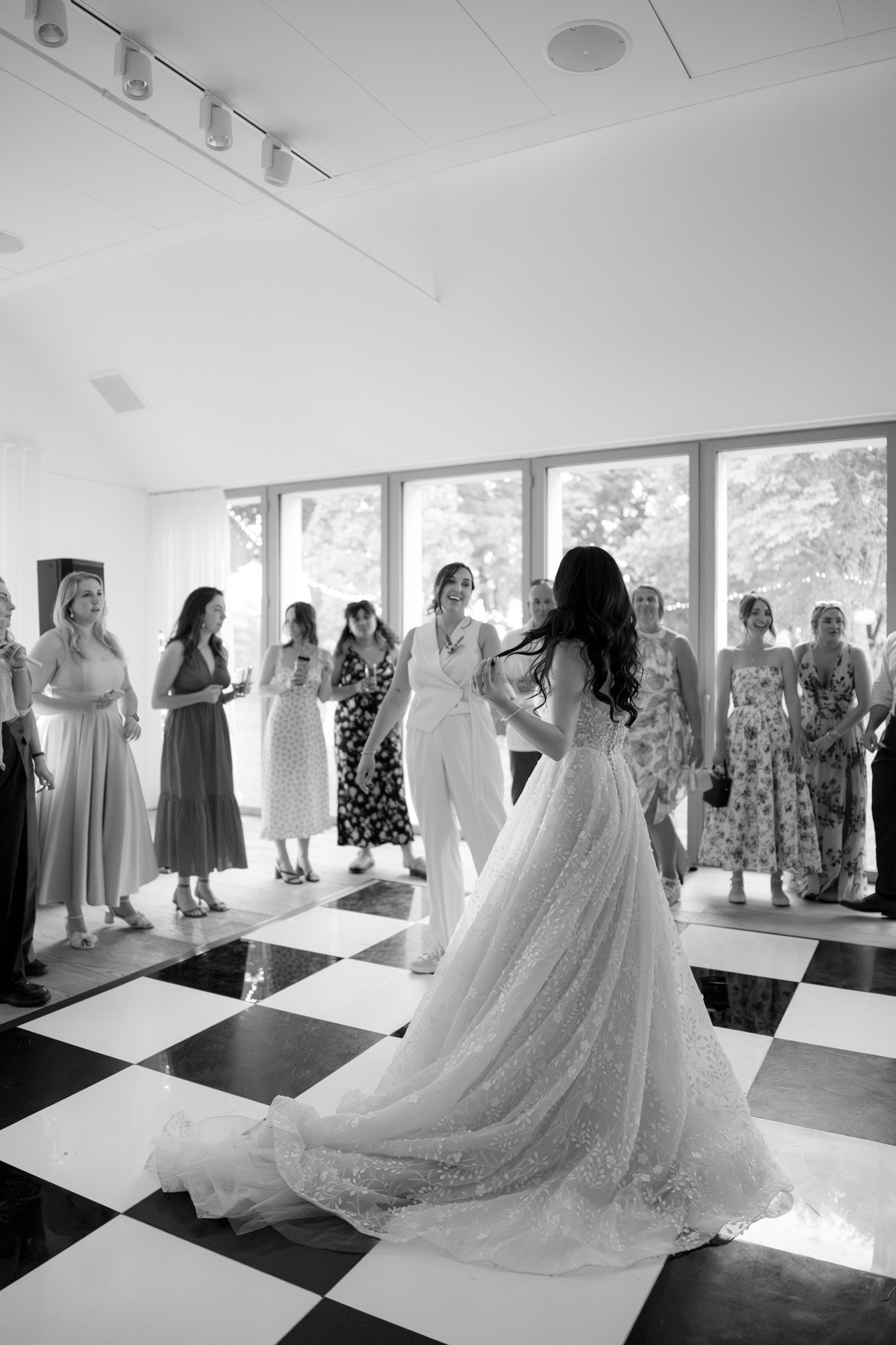 A bride in a long gown dances on a checkered floor surrounded by guests in dresses, inside a bright room with large windows. Spring Dordogne Wedding Photographer