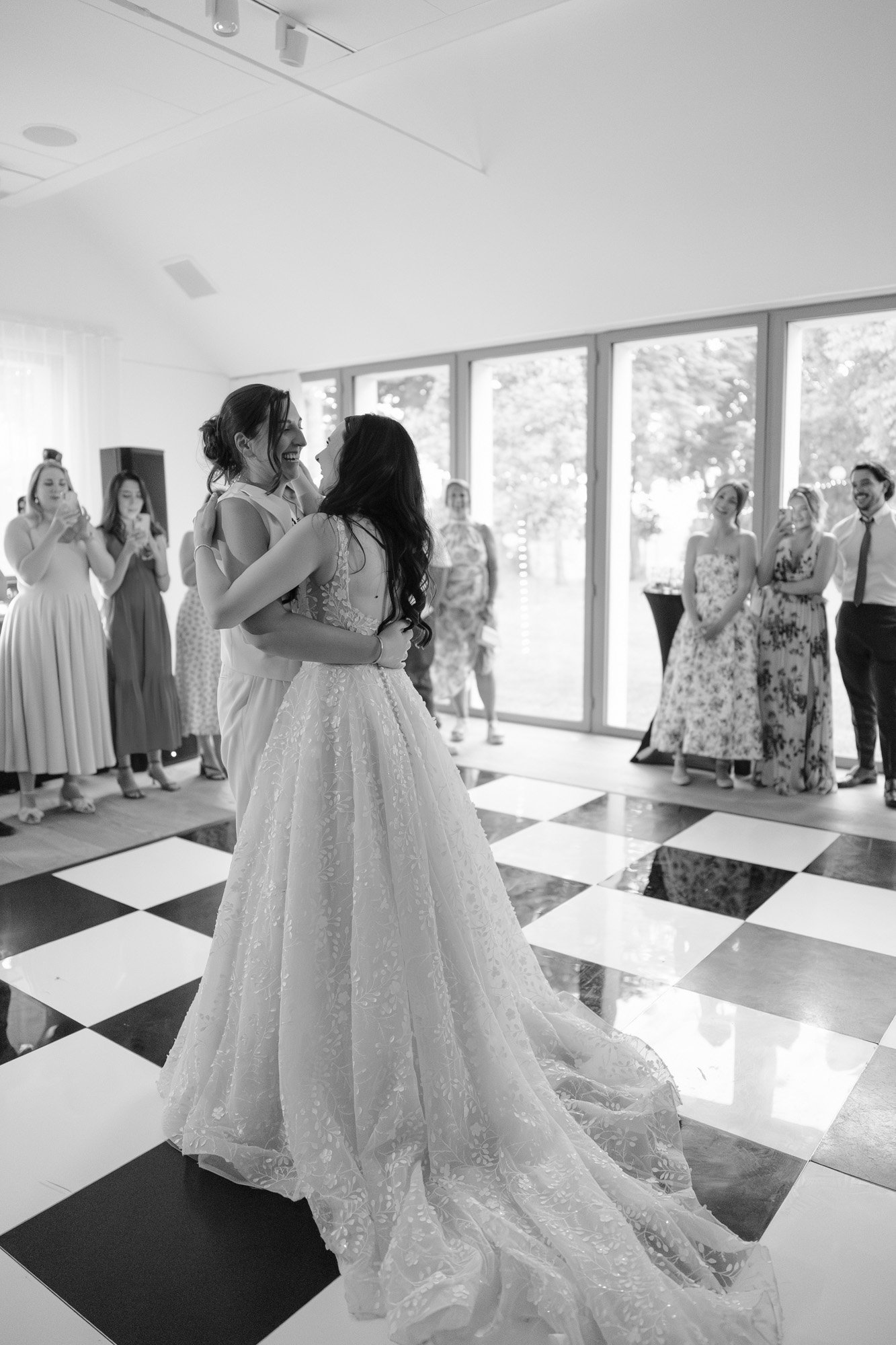 Two women in wedding dresses dance together on a checkered floor while guests watch and smile in a brightly lit room. Spring Dordogne Wedding Photographer