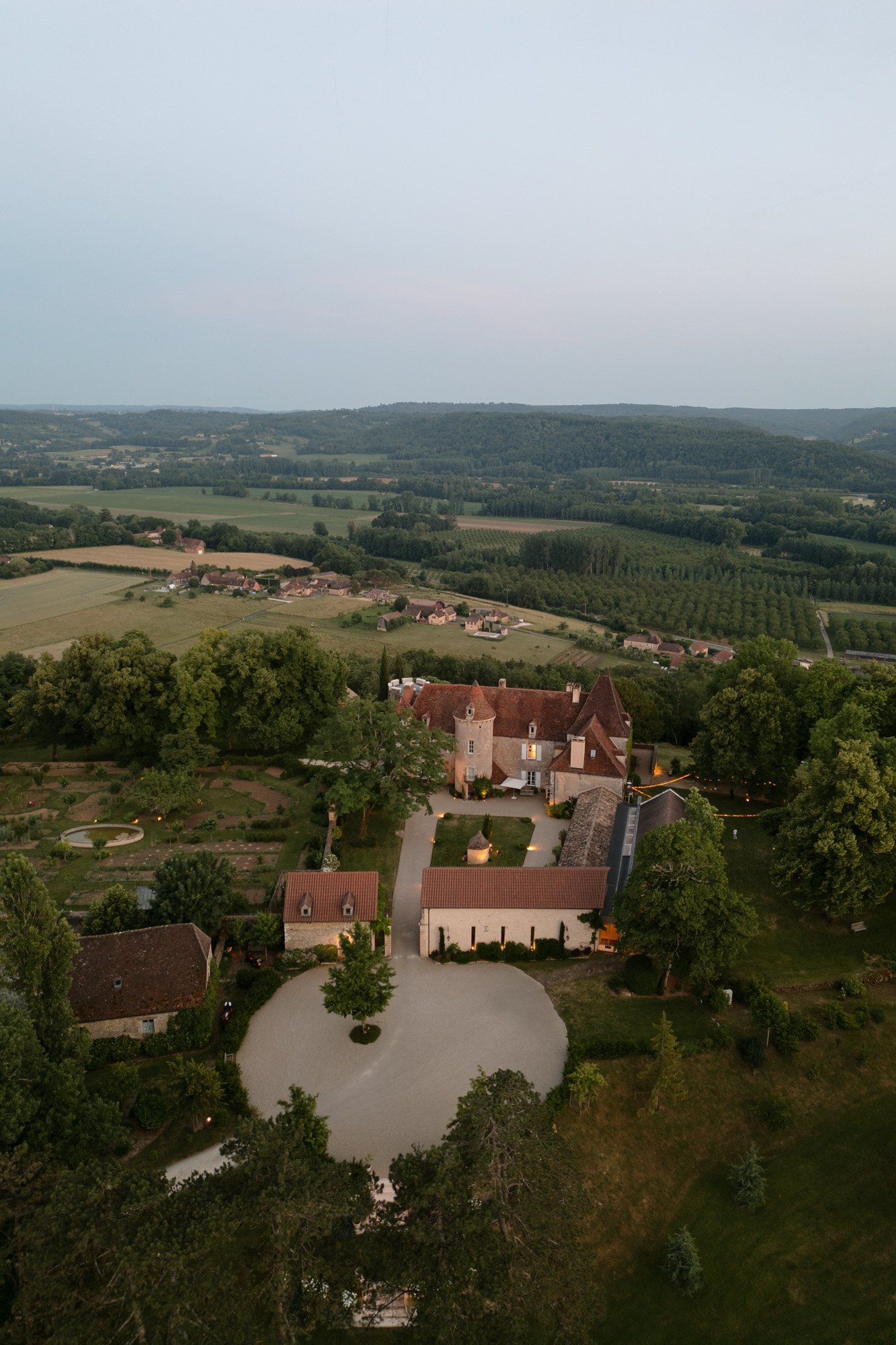 Aerial view of a countryside estate with several buildings, circular driveway, gardens, and surrounding fields, under a clear sky. Spring Dordogne Wedding Photographer
