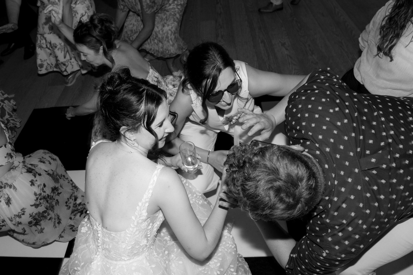 A group of people in formal attire kneel and laugh together on a checkered dance floor at an indoor event.