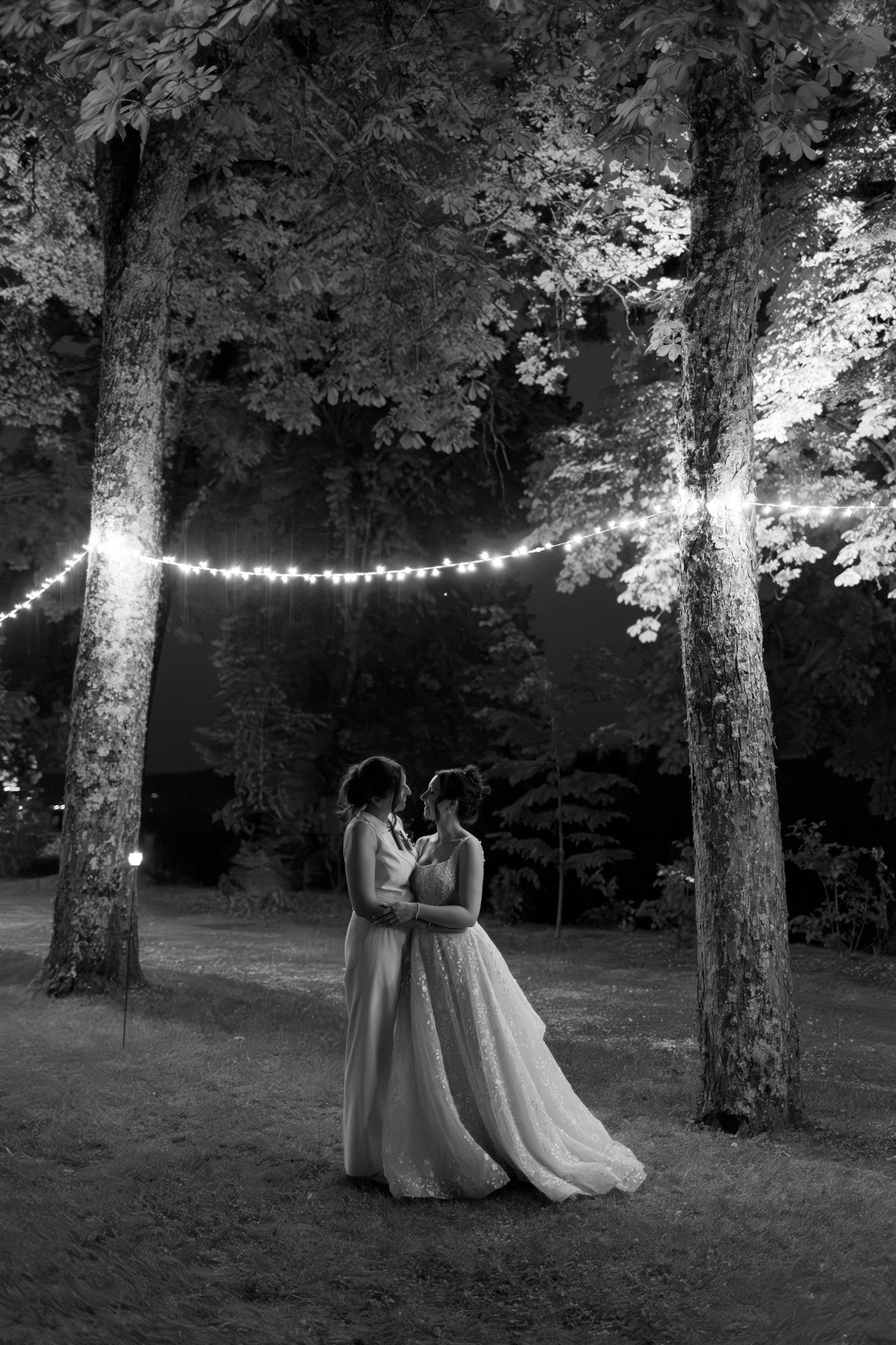 Two women in formal dresses stand close together under string lights between trees at night, in a grassy outdoor setting. Spring Dordogne Wedding Photographer