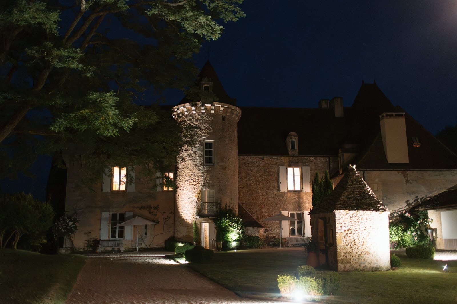 A stone chateau with a round tower and pointed roofs is illuminated at night, surrounded by manicured lawns, trees, and small outbuildings. Spring Dordogne Wedding Photographer
