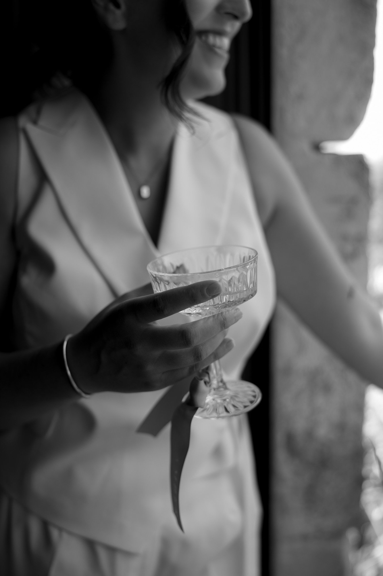 A person in a sleeveless outfit holds a crystal glass of beverage near a window, smiling. The image is in black and white.