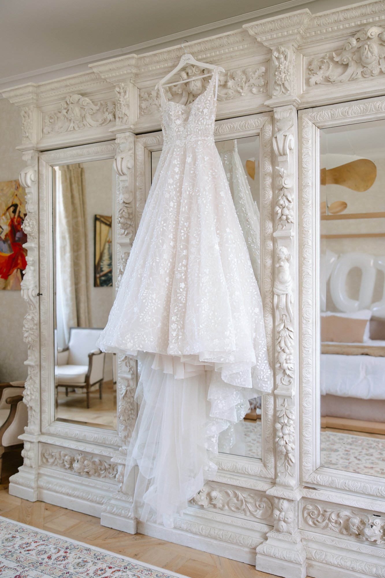 A white, floral-patterned wedding dress hangs on an ornate mirrored wardrobe in a well-lit, elegant room.